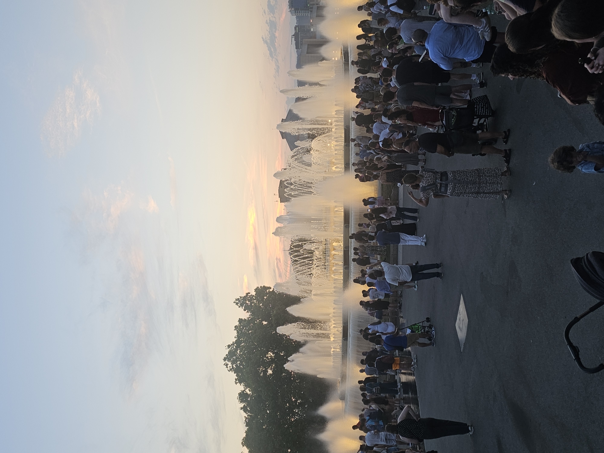 The image depicts a large crowd of people gathered around a multi-tiered fountain with water jets spraying upwards. The fountain is illuminated and appears to be part of a public square or park. The time of day is sunset, with a warm, soft light casting a glow on the sky and the water. The sky is a mix of soft blues and oranges, with some wispy clouds. In the background, there are buildings and trees, suggesting an urban setting.
The people in the foreground and midground are diverse, with many individuals and families present. They are dressed in casual summer clothing, and their postures suggest they are observing the fountain display. Some are holding up phones, possibly to take pictures or videos. The overall mood seems to be one of enjoyment and shared experience.
Notable details include the detailed architecture of the fountain structure, the spray of the water creating a mist, and the reflections of the light on the wet pavement around the fountain. The presence of a stroller suggests that children are among the attendees. The location is identified as Barcelona, Spain. No visible text is present in the image.