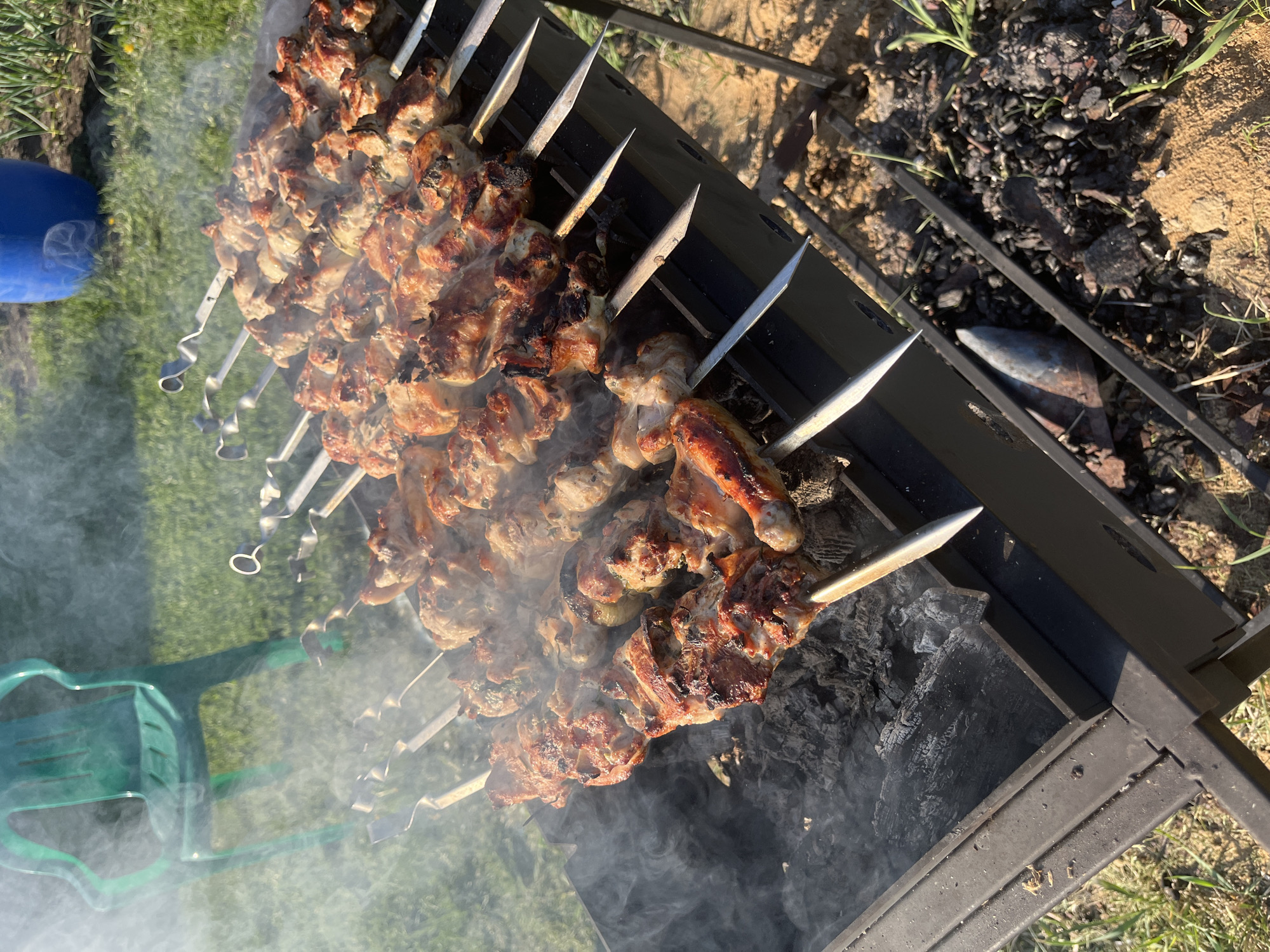 The image shows a barbecue grill with multiple metal skewers laden with grilling meat, likely shashlik. The skewers are arranged across the top of the grill, which appears to be charcoal-fueled, judging by the smoldering coals underneath and the rising smoke. The surrounding environment consists of grass and some sandy, bare earth. A blue container and a green chair are visible in the periphery. The location is identified as Сапуголи, Russia.
