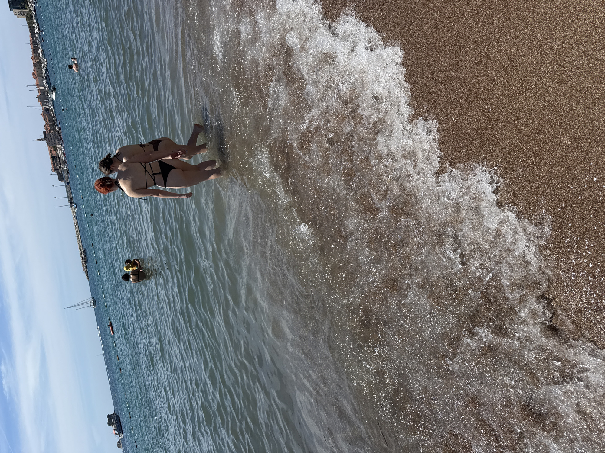 Two individuals, identified as women by their attire and physiques, are walking into the sea from a beach in Budva, Montenegro. They are both wearing bikinis. The woman on the left has red hair and is wearing a black bikini. The woman on the right is wearing a dark brown bikini. They are walking side-by-side, with their hands clasped.
The beach appears to be sandy, with small pebbles mixed in. The sea is a clear, blue-green color, with gentle waves lapping at the shore. In the background, several boats and yachts are visible on the water. A city skyline, characterized by red-tiled roofs and buildings, is situated along the coast. The sky is bright blue with scattered clouds, indicating a sunny day. Other beachgoers are present in the water and on the shore, though they are not the primary focus of the image. The overall scene depicts a typical summer day at the beach in Budva.