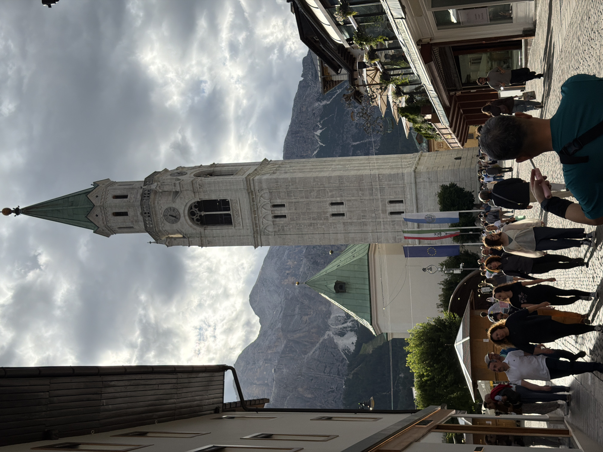 The image depicts a street scene in a European-style town, likely in the Alps, given the presence of a tall bell tower and rugged mountains in the background. The bell tower, made of pale stone with green accents, is a prominent feature in the center of the frame. A smaller building with a green, conical roof is situated in front of it. The street is paved with cobblestones and lined with buildings, some of which appear to be shops or cafes with awnings and outdoor seating. A crowd of people is walking along the street, some dressed casually, others in more formal attire. The sky is cloudy, with dramatic shafts of sunlight breaking through, suggesting either the beginning or end of the day. The overall atmosphere is one of a bustling, picturesque town square.