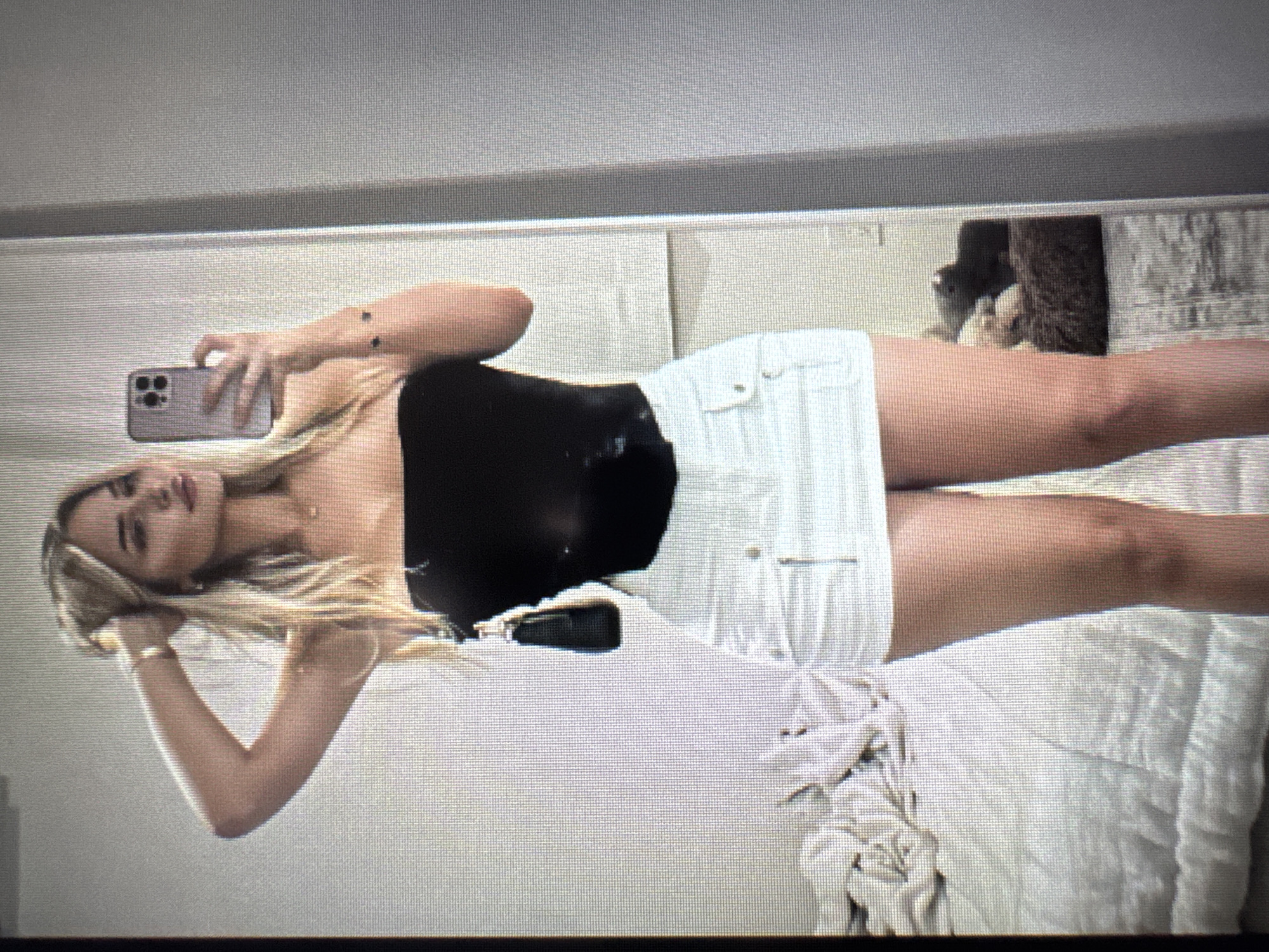 A young woman with long, straight blonde hair is depicted taking a mirror selfie in an indoor setting. She is positioned to the left of the frame, facing slightly away from the camera, with her head tilted and looking towards her reflection. She is wearing a black strapless top and white shorts. A gold-toned bracelet is visible on her left wrist.
Her right hand holds a light-colored smartphone, featuring three distinct camera lenses, pointed towards the mirror to capture the image. Her left arm is bent, with her hand resting near her head, lightly touching her hair. Her expression is neutral and composed.
The background, seen through the mirror's reflection, consists primarily of light-colored, possibly white or off-white, surfaces, likely bedding or furniture. To the right, a dark, textured object, possibly a stuffed animal or decorative item, is partially visible. The overall scene suggests a bedroom or personal living space. The image itself exhibits a noticeable pixelated or screen-door effect, indicating it may be a photograph of a digital screen rather than an original digital file. No discernible text is visible.