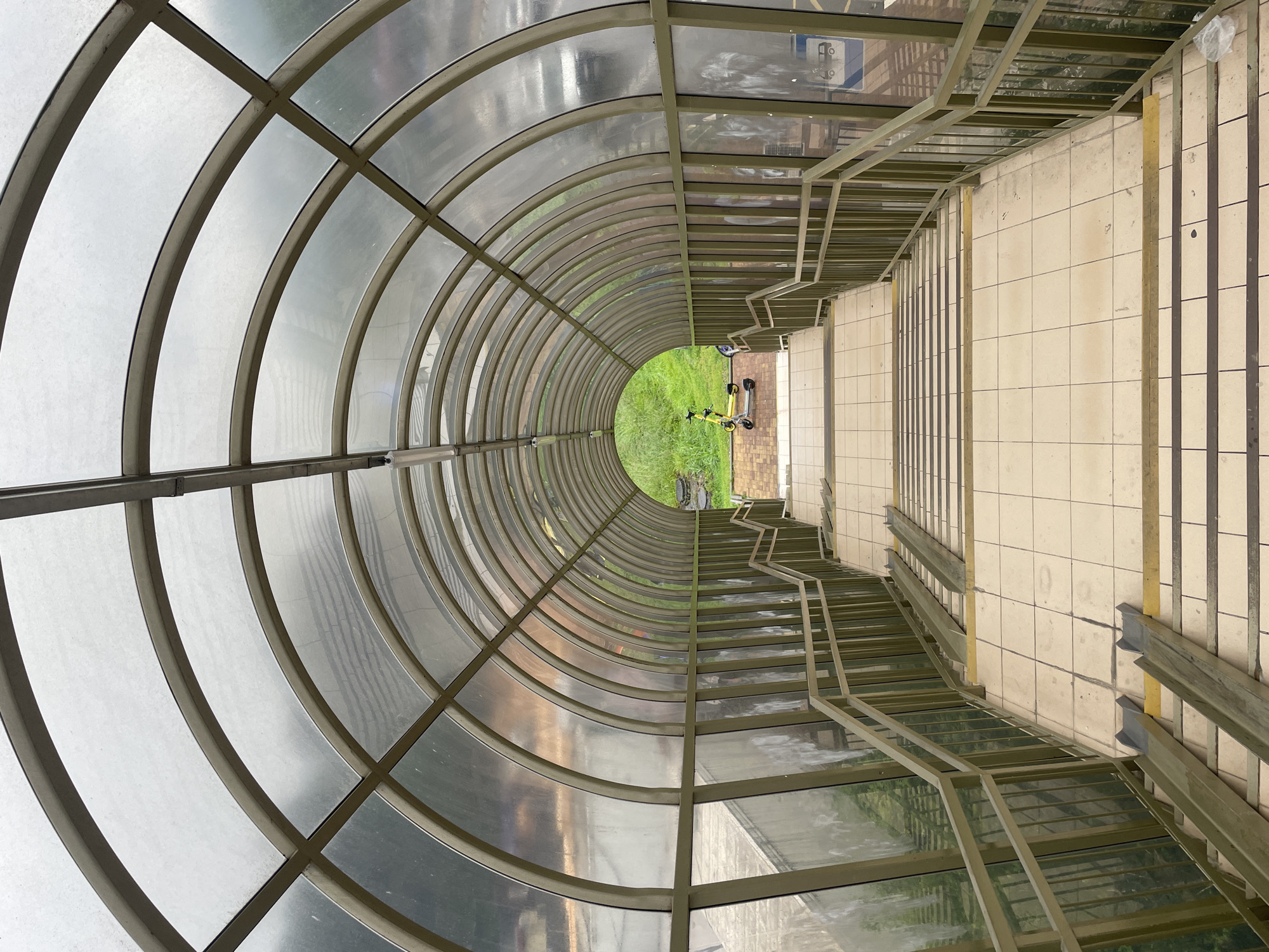The image shows the interior of a covered pedestrian walkway, potentially an underpass or overpass. The walkway has a semi-circular transparent roof composed of arched metal frames with transparent or translucent panels. On the right, there are stairs leading downwards, with metal railings along the sides. The floor appears to be tiled. At the far end of the walkway, an opening reveals a green, grassy area with two parked scooters.
The image is associated with the city of Sochi, Russia.