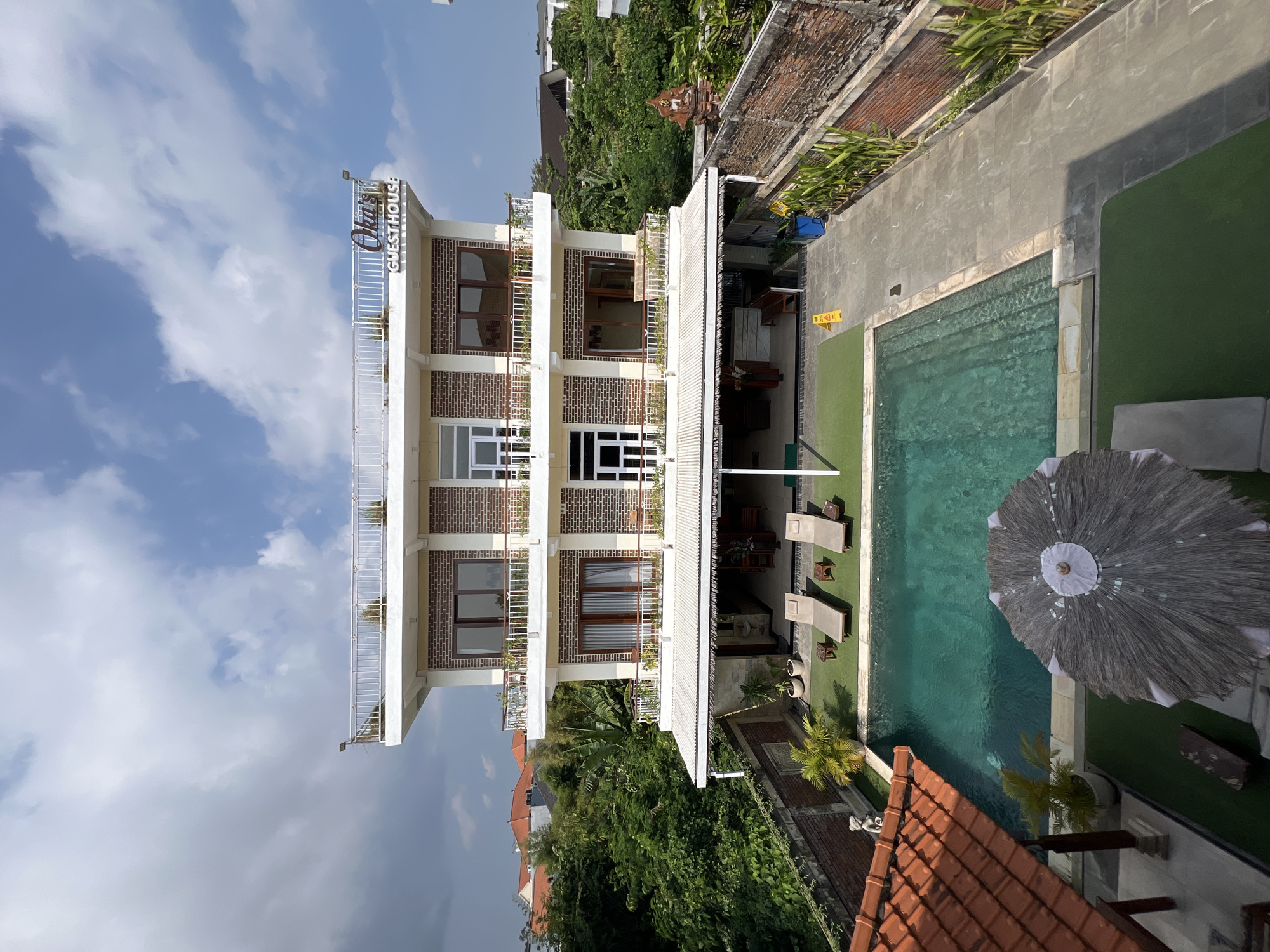 The image shows a multi-story guesthouse with a swimming pool in the foreground, located in North Kuta, Indonesia. The guesthouse is a modern building with a brick facade and multiple balconies. A sign on the top floor reads "Oka's Guesthouse". The swimming pool is turquoise and surrounded by lounge chairs. There is lush green vegetation around the property, including trees and plants. The sky is partly cloudy. No people are visible in the image.