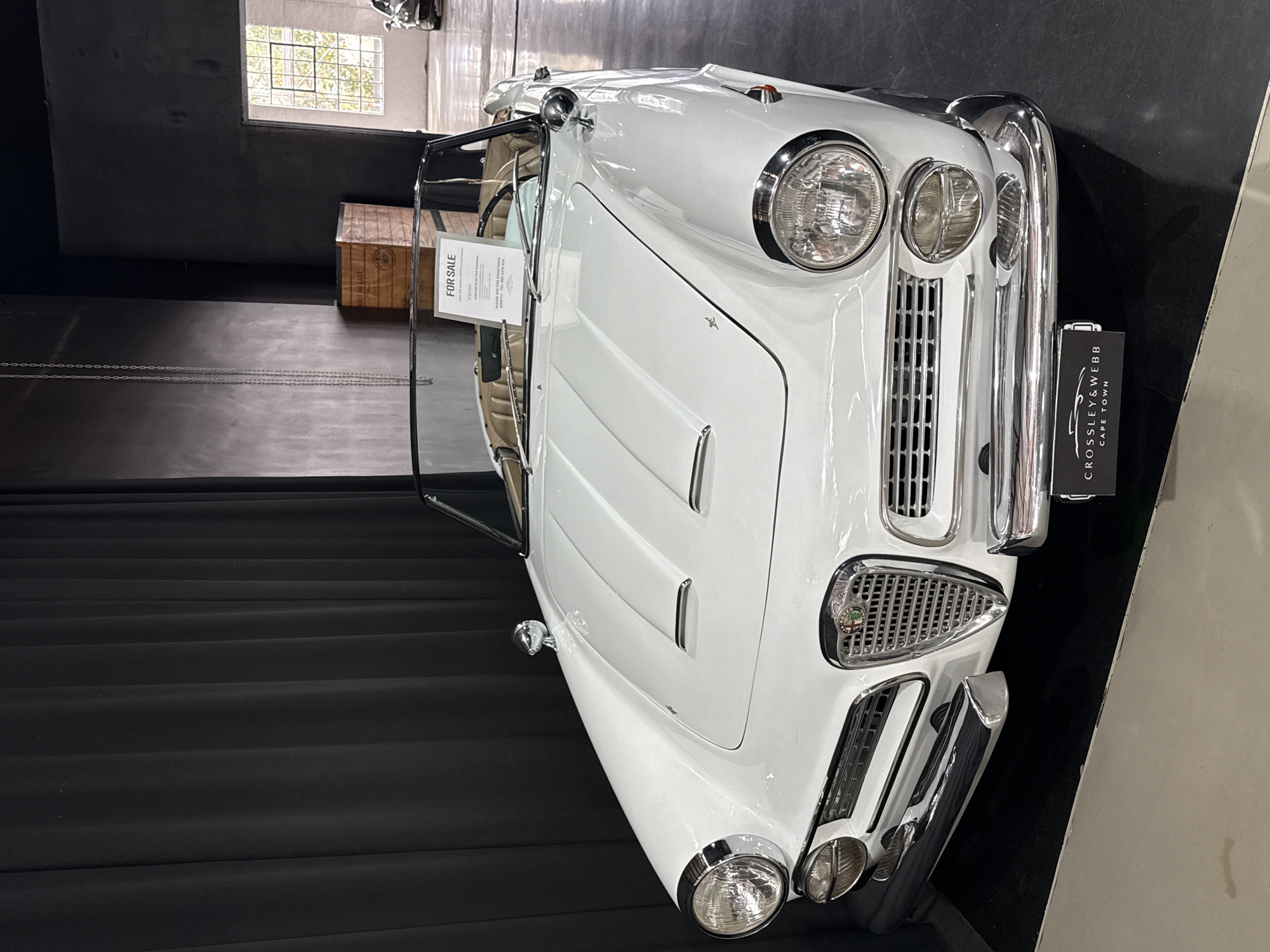 The media file captures a pristine white classic convertible car, likely from the mid-20th century, displayed indoors within what appears to be a showroom or museum. The car is positioned diagonally, its front facing towards the upper right of the frame, with the image itself rotated 90 degrees. It showcases numerous chrome accents, including dual headlights on each side, a prominent vertical-slat grille, bumper bars, side mirrors, and hood vents. The visible interior features a light tan or beige color.
The setting is characterized by dark, possibly black, walls and a dark, ridged floor, which provides a stark contrast to the white vehicle. Natural light streams in from a window visible in the upper left background, revealing a grid pattern and some green foliage outside, indicating it is daytime. A wooden crate partially visible in the background is labeled "BUILT TO LAST".
A white "FOR SALE" sign is attached to the car's windshield frame, with text stating "PLEASE ASK FOR" and contact information: "GARETH TEL: 083 5370 456". A prominent black sign positioned beside the car's front bumper displays "CROSSLEY & WEBB" and "CAPE TOWN", confirming the location as Cape Town, South Africa. No individuals are present in the image; the car is stationary, on display for potential buyers. The overall presentation emphasizes the vehicle's elegance and historical appeal.