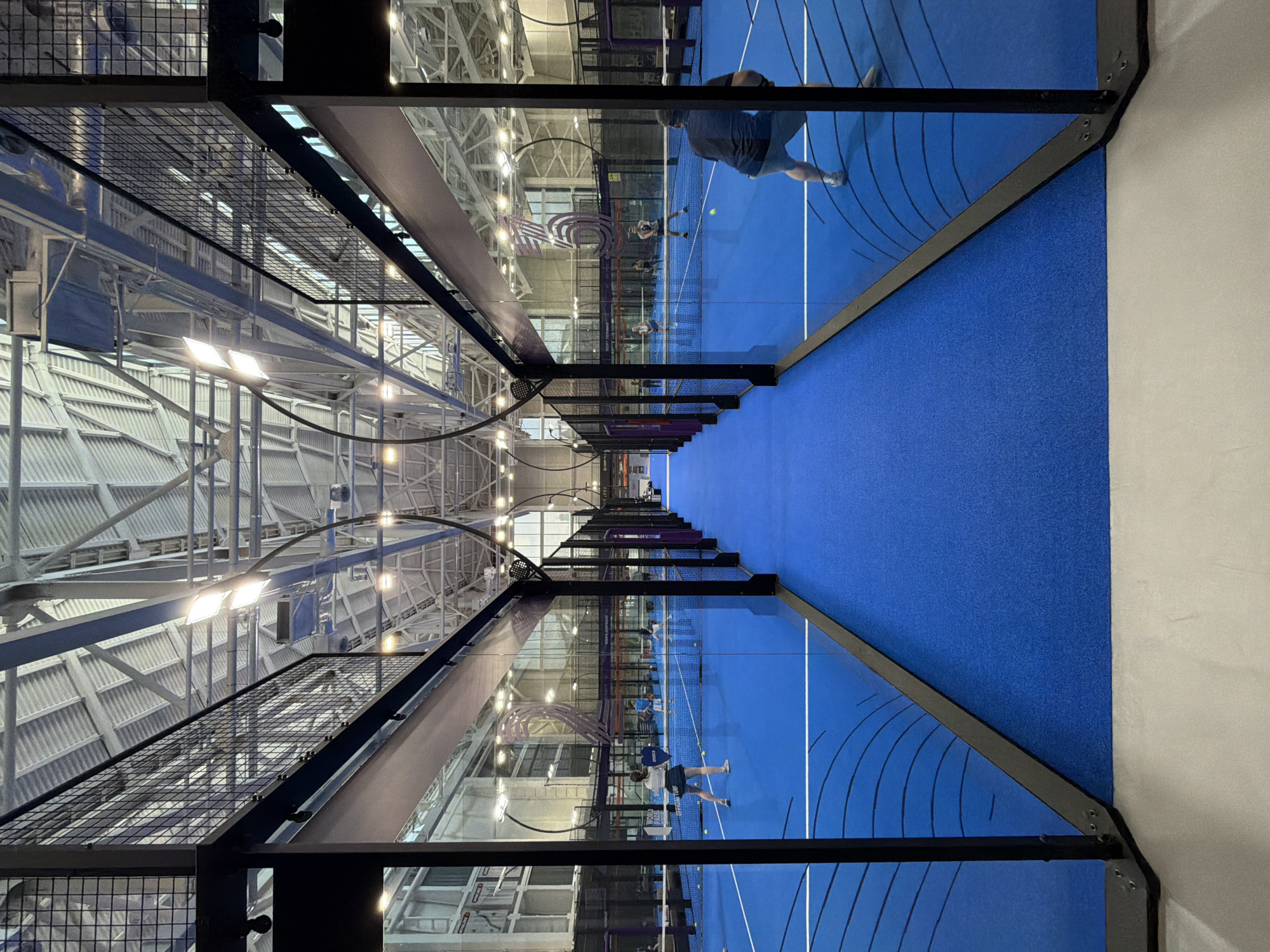 The image captures the interior of what appears to be a padel tennis facility, potentially in Moscow, Russia, given the request. The scene is oriented in a way that the padel court extends from bottom to top, framed by the structure of the court itself, including the glass and metal framework. Players are visible on the blue court, engaged in the game. The roof structure is high and industrial, featuring metal supports and lighting fixtures. The overall environment is well-lit. The walls of the facility are light-colored, contrasting with the vibrant blue of the court.