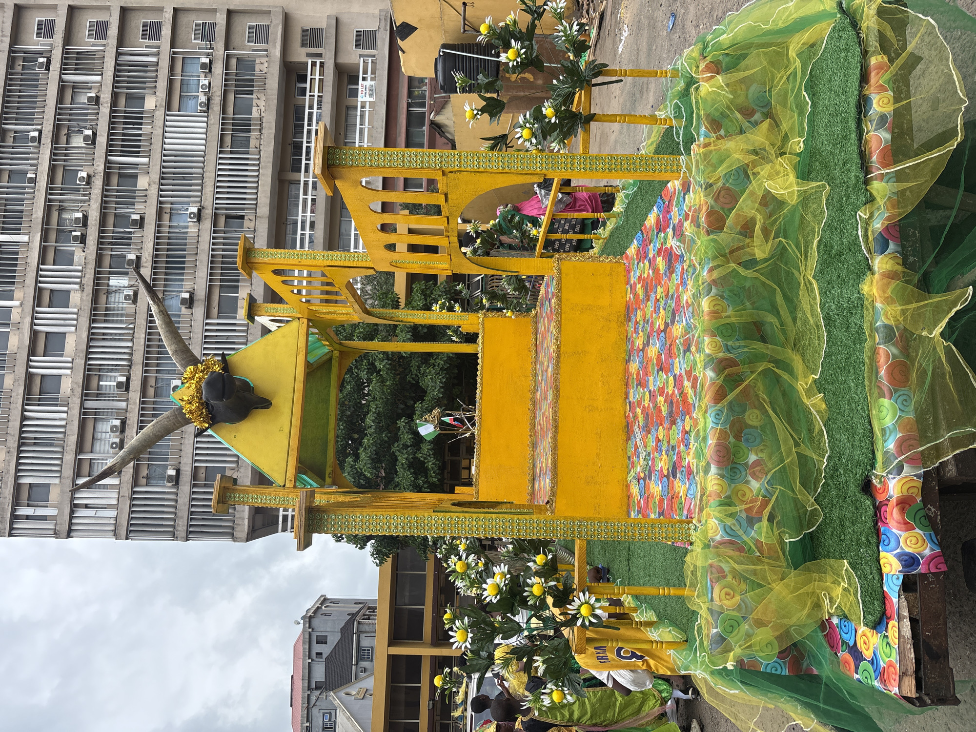 The image shows a street parade float in Lagos, Nigeria. The float is predominantly yellow, with a decorative bull skull mounted on its front. It's adorned with artificial flowers and swaths of green and yellow tulle. The platform is covered with a colorful, swirled pattern and artificial turf. In the background are buildings, a multi-story office block with visible air conditioning units and a smaller residential structure. People are visible around the base of the float, though their faces are not distinct. The sky is overcast.