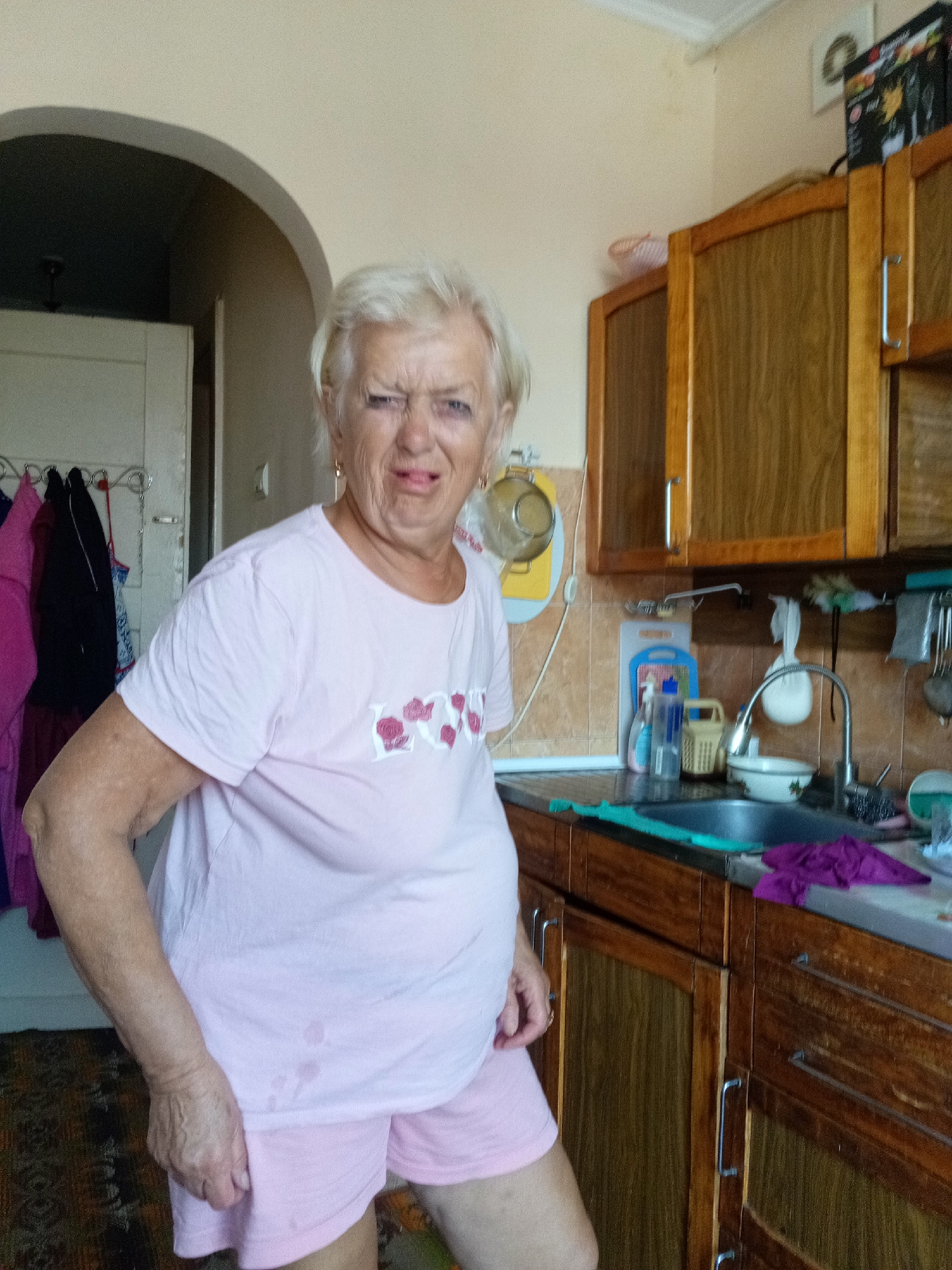 The image shows an elderly woman standing in a kitchen. She has white hair and is wearing a light pink t-shirt and pink shorts. Her face is contorted in an expression of displeasure, with her eyebrows furrowed and her mouth slightly open. Behind her, wooden cabinets line the walls, and a sink with a faucet is visible on the counter. Various kitchen items, including bottles, a bowl, and cleaning supplies, are scattered around the sink. A rack with some clothing items hangs on the wall to the left of the woman. The overall impression is of a domestic interior, possibly in an older building, with an unkempt appearance. The location is identified as Tiachiv, Ukraine.