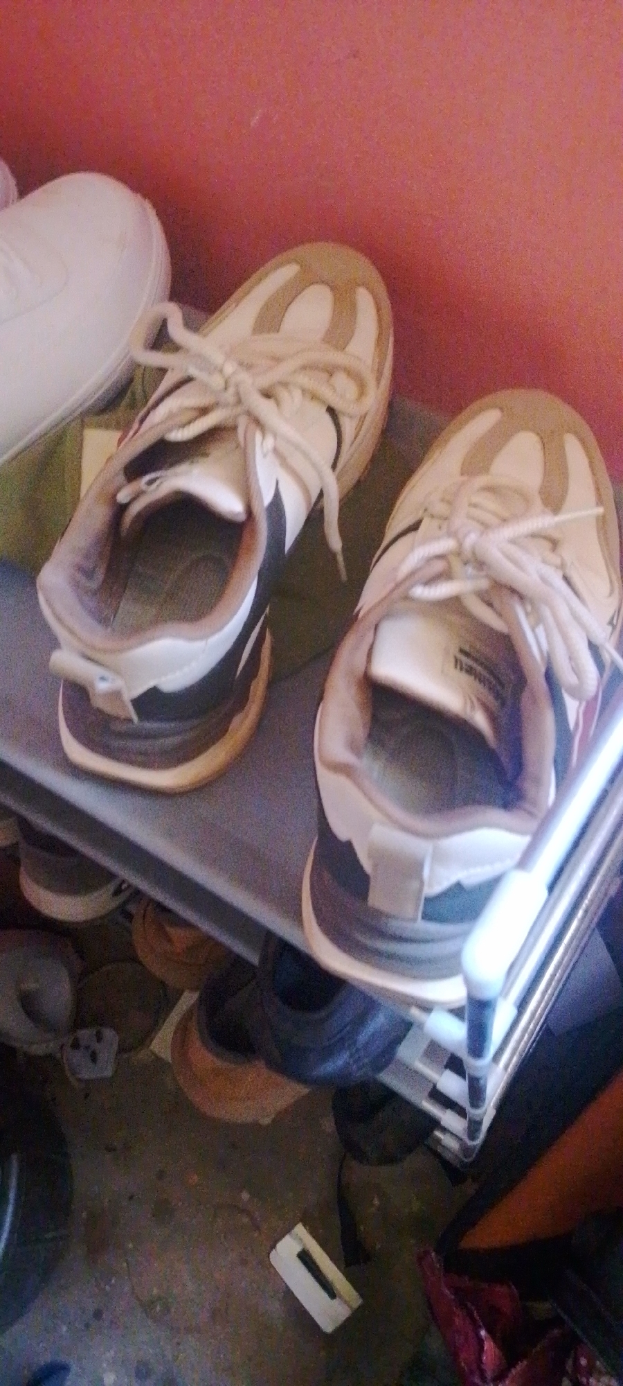A pair of white and tan sneakers with tan laces are displayed on a gray shoe rack. One sneaker is placed inside the other. The shoe rack is located on a dusty concrete floor. Several other shoes are visible on lower shelves and the floor. A pinkish-red wall is in the background. The scene appears to be indoors, possibly in a closet or storage area.