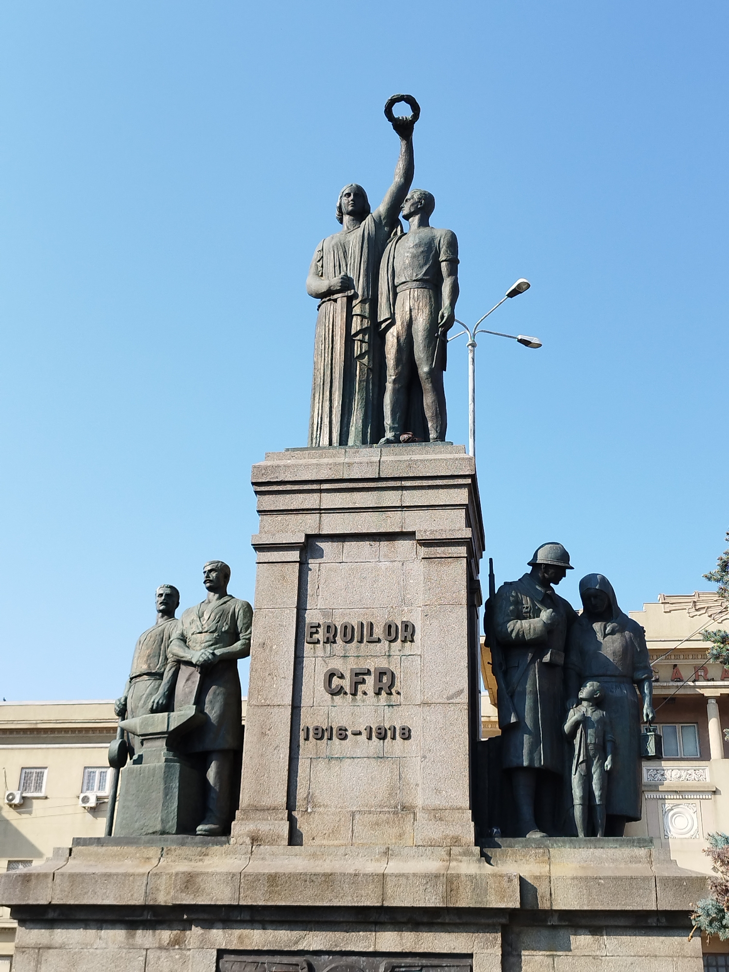 The image shows a large, multi-figure bronze monument situated in a public space, likely a square, in Bucharest, Romania. The monument is dedicated to the heroes of the CFR (Căile Ferate Române, Romanian Railways) and commemorates the years 1916-1918.
The central element of the monument is a tall stone pedestal with inscriptions. On top of the pedestal stand two prominent figures. The figure on the left is draped in a toga-like garment and holds a laurel wreath aloft with their right arm. The figure on the right, dressed in working-class attire, stands beside the first figure, looking upward.
Flanking the central pedestal are other bronze figures. To the left, two men are depicted. One stands behind an anvil, holding a hammer, suggesting a blacksmith or a laborer. The other man stands beside him, looking forward. To the right of the central pedestal, a group of three figures is present: a man in military uniform holding a rifle, a woman in a headscarf, and a child looking up at them.
The monument is set against a clear blue sky. In the background, a building with multiple windows and air conditioning units is visible, along with trees and streetlights. The overall scene depicts a solemn tribute to fallen heroes, rendered in a realistic style.