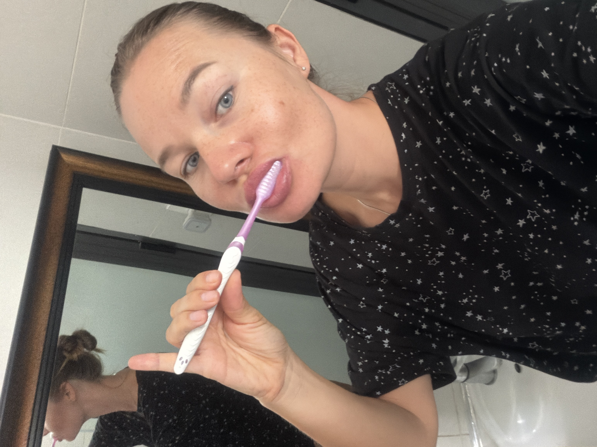 A woman with light skin, blue eyes, and freckles is brushing her teeth in a bathroom. She has her hair pulled up in a bun. She is wearing a black t-shirt with white stars. The toothbrush is white with a purple tip. A mirror behind her reflects her side profile. The bathroom appears to be tiled with white tiles. There is a sink in the foreground. The image does not provide information about the city of Lâm Viên - Đà Lạt, Vietnam.