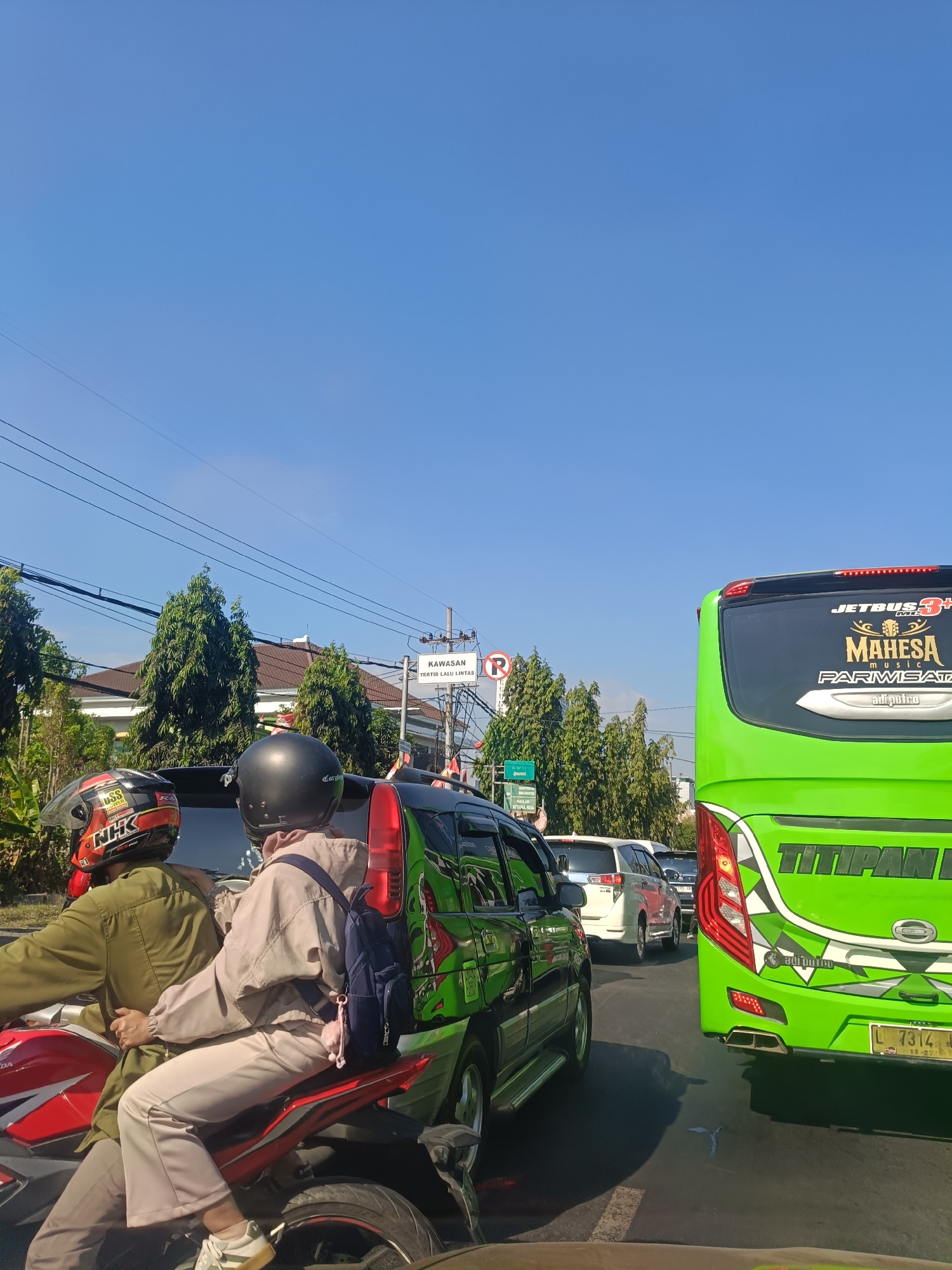 The image shows a traffic jam on a sunny day in Dahanrejo, Indonesia. In the foreground, two people are riding a red motorcycle. The person in the back is wearing a beige jacket and a blue backpack. The person in the front is wearing a green jacket and a red and black helmet. The motorcycle is stopped behind a black SUV, which has a green and black design on its side. To the right of the SUV is a bright green bus with "TITIPAN BUS" written on the back. The bus has a license plate that reads "L 7314 JK". In the background, there are several other cars and trees. A street sign with "KAWASAN TERTIB LALU LINTAS" is visible on the right side of the image. The sky is clear and blue.