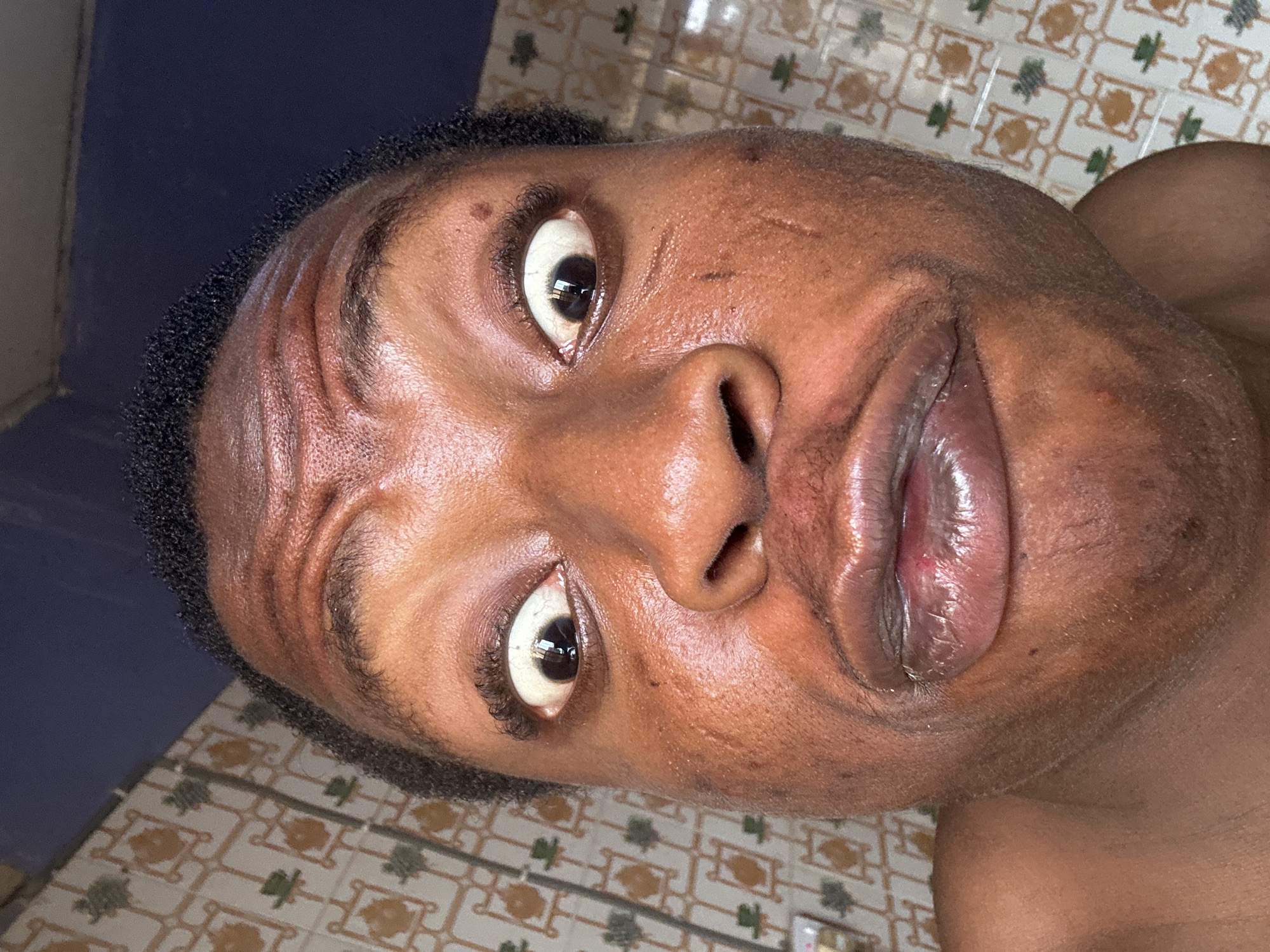A close-up portrait of a dark-skinned individual with short, dark hair. The person is looking directly at the camera with wide eyes. The background includes a wall and patterned floor tiles. The location is Unknown, Unknown.