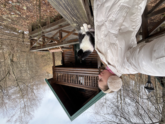 The image captures an outdoor scene in Crkvine, Montenegro, featuring an adult female and a domestic cat on a wooden deck in front of a log cabin. The female, positioned on the left side of the frame, is seen in profile, facing left, with her left arm extended, holding an unseen device consistent with taking a self-portrait. She wears a light-colored, likely beige or cream, shiny puffer jacket with a fleece-lined collar, a pink garment visible underneath, and large, fluffy beige earmuffs. Her light-colored hair is partially visible.
To her right, a black and white domestic cat sits upright on a sturdy wooden railing, facing right, with its head turned away from the camera. The cat exhibits a white chest, paws, and facial markings against a primarily black body.
In the background, a two-story wooden log cabin with a dark green A-frame roof is prominently featured. The cabin has a second-floor balcony with a wooden railing and a first-floor deck where a wooden bench is visible. The structure is constructed from dark brown logs. Surrounding the cabin and subjects is a dense forest of deciduous trees with bare branches, suggesting late autumn or winter. The ground is covered with fallen brown and orange leaves. An old-fashioned black outdoor lamp post is visible in the upper left portion of the image, behind the female. The sky is uniformly overcast and light grey.