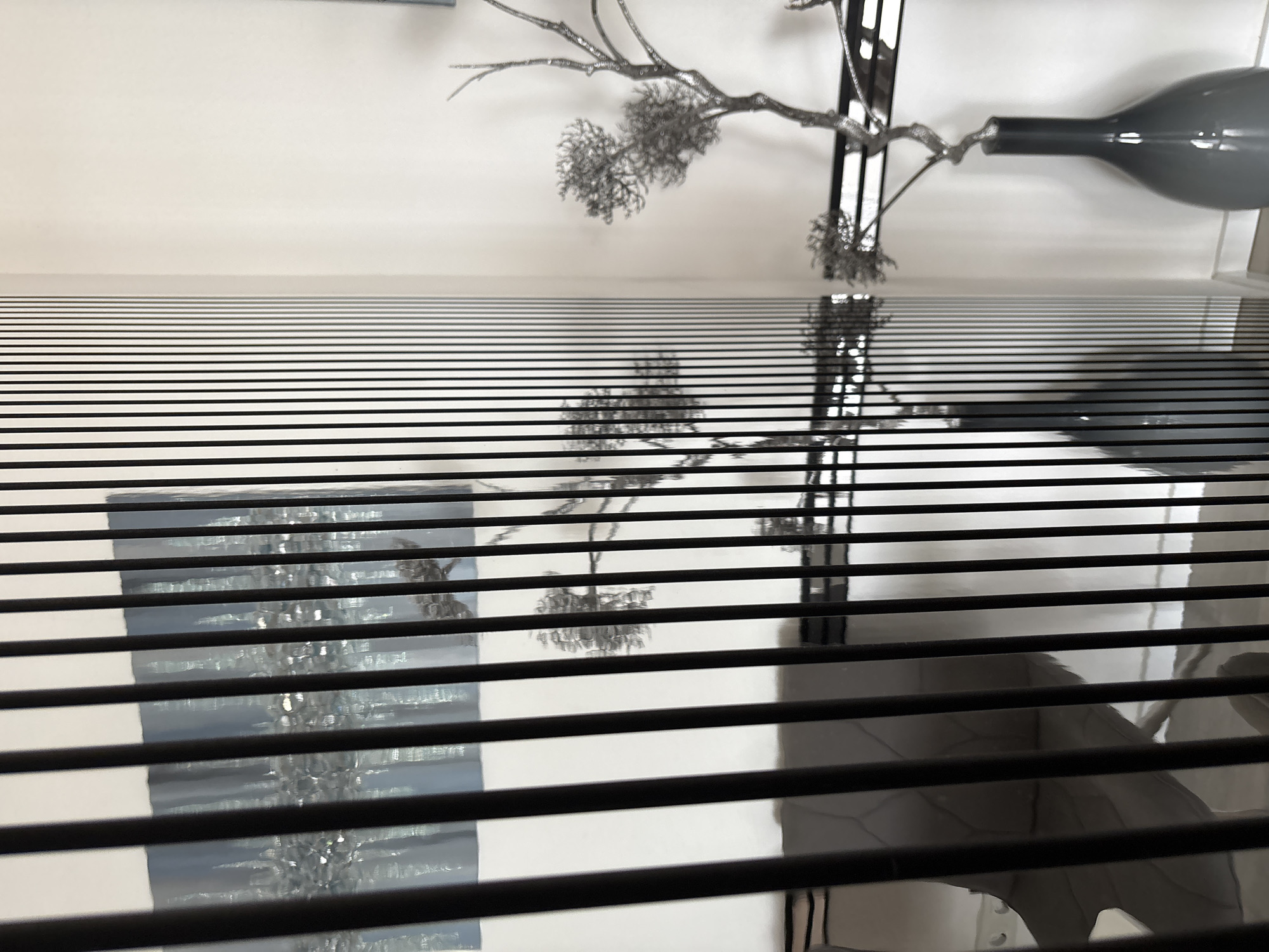 The image shows a modern interior with a focus on decorative elements and a partition. A tall, dark grey vase with a metallic, bare tree branch is positioned to the right of the frame, set against a plain white wall. The partition, which dominates the left and center of the image, is composed of thin, vertical black slats. This partition creates a visual barrier and reflects the environment behind it. Through the slats, a portion of a grey upholstered chair is visible on the left. Behind the partition and partially obscured by it is a wall-mounted artwork featuring abstract, vertical strokes of blue and shimmering silver, resembling a reflective landscape or water scene. The overall aesthetic is minimalist and sophisticated.