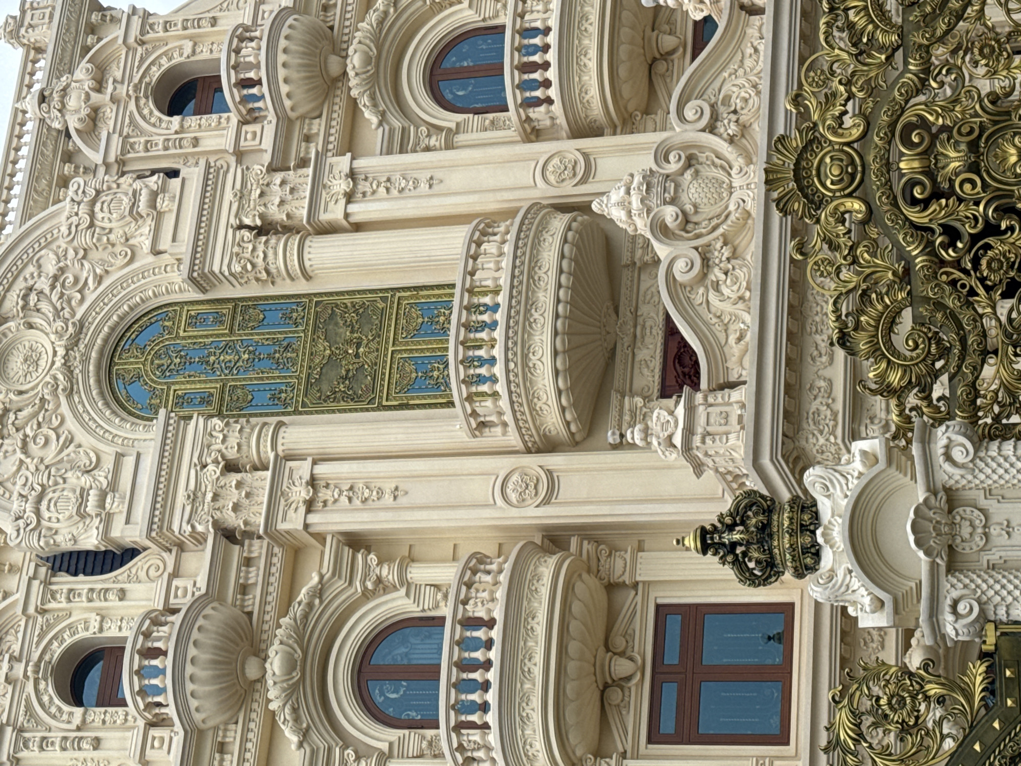 The image shows the ornate facade of a building, likely a historical or grand residence. The architecture is characterized by intricate carvings, elaborate decorative moldings, and multiple balconies with ornate railings. The building's color is a pale cream, contrasting with the dark wooden frames of the windows and the decorative gold elements, such as the gate in the foreground and a crown-like finial on the building. The central section of the building features a large, decorative window with blue glass and gold detailing. The overall impression is one of opulence and historical grandeur. The building is located in Da Lat, Vietnam.