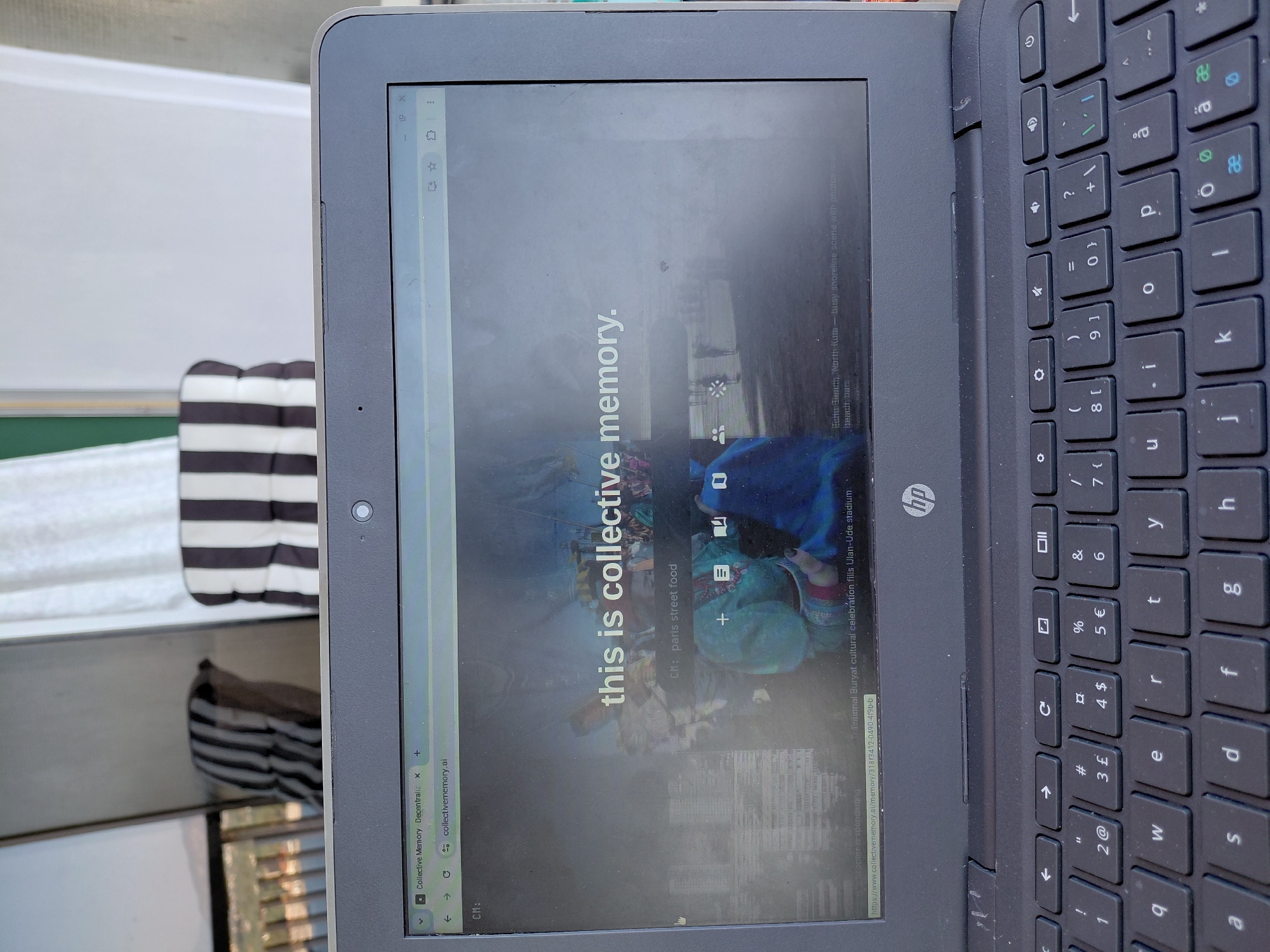 A laptop is open, displaying a website with text overlayed on an image. The text reads "this is collective memory." Below this, it says "CM: paris street food." The website address visible in the browser bar is "collectivememory.ai." The lower part of the image shows a portion of the laptop's keyboard and trackpad. The background shows a blurred view of furniture and possibly a balcony or outdoor space. There are no discernible elements in the image that can be used to determine its location within the city of Municipal Unit of Aegio, Greece.
