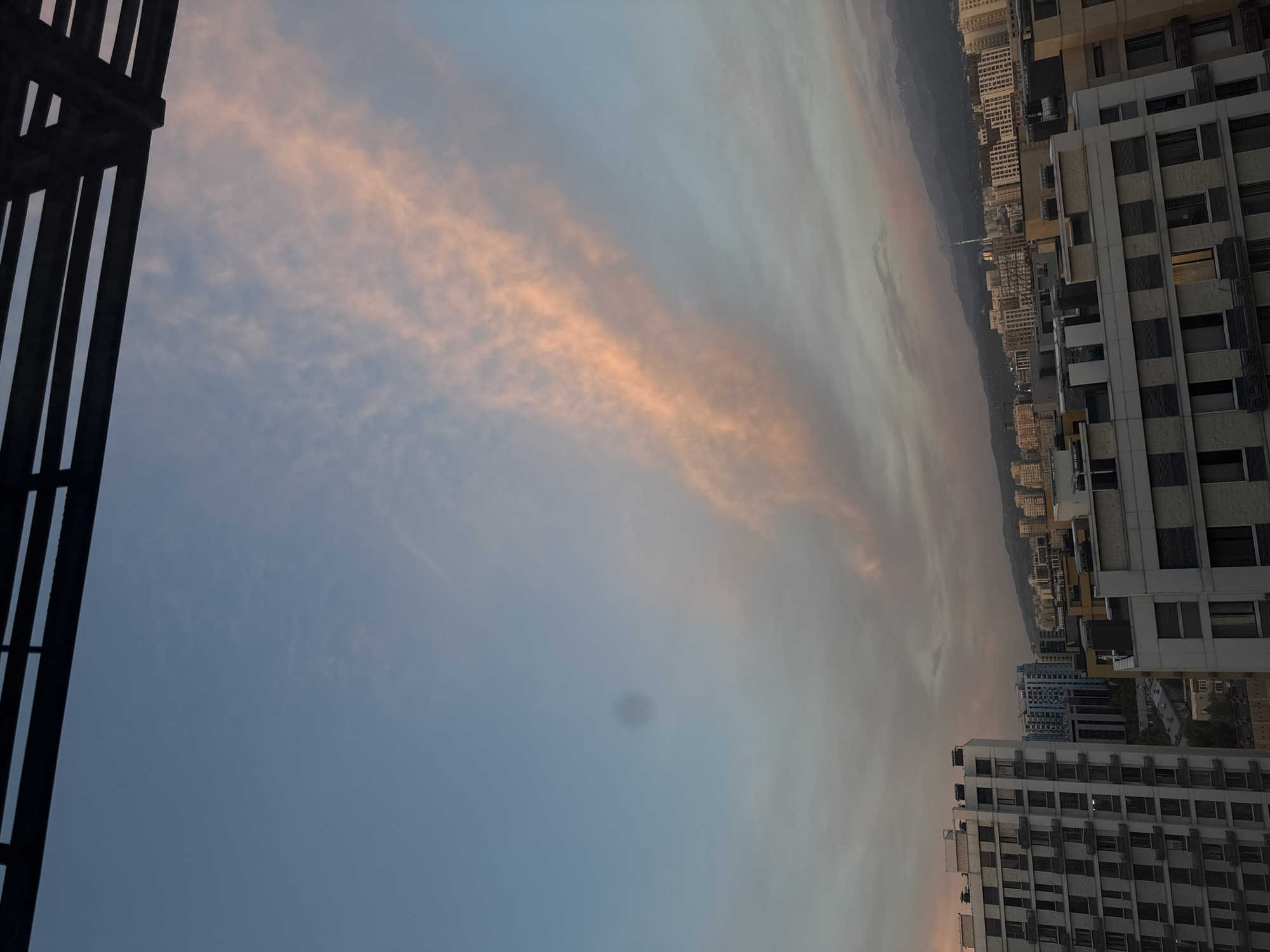 The image captures a cityscape at dusk, likely in Almaty, Kazakhstan, given the presence of mountains in the background. The foreground features a dark, abstract metallic structure, possibly a balcony railing or part of a building's facade, on the left side of the frame. The majority of the image is dedicated to the sky, which is transitioning from day to night. Wispy clouds are visible, tinged with hues of peach and pink, suggesting the warm glow of a setting sun.
Beyond the immediate foreground, a dense urban landscape unfolds. Numerous buildings of varying heights and architectural styles populate the scene, stretching towards the distant mountains. The buildings are predominantly light-colored, with visible windows and balconies. Some buildings appear to be residential blocks, while others might be commercial structures. The overall impression is of a sprawling city nestled against a natural backdrop.
The time of day is clearly evening or twilight, as indicated by the soft lighting and the colors in the sky. The weather appears clear, with only scattered clouds. There are no people or animals visible in the image. The mood is tranquil and serene, evoking a sense of calm as the day ends. The presence of the mountain range provides a strong geographical cue, suggesting a location with significant elevation changes. No discernible text is present in the image.