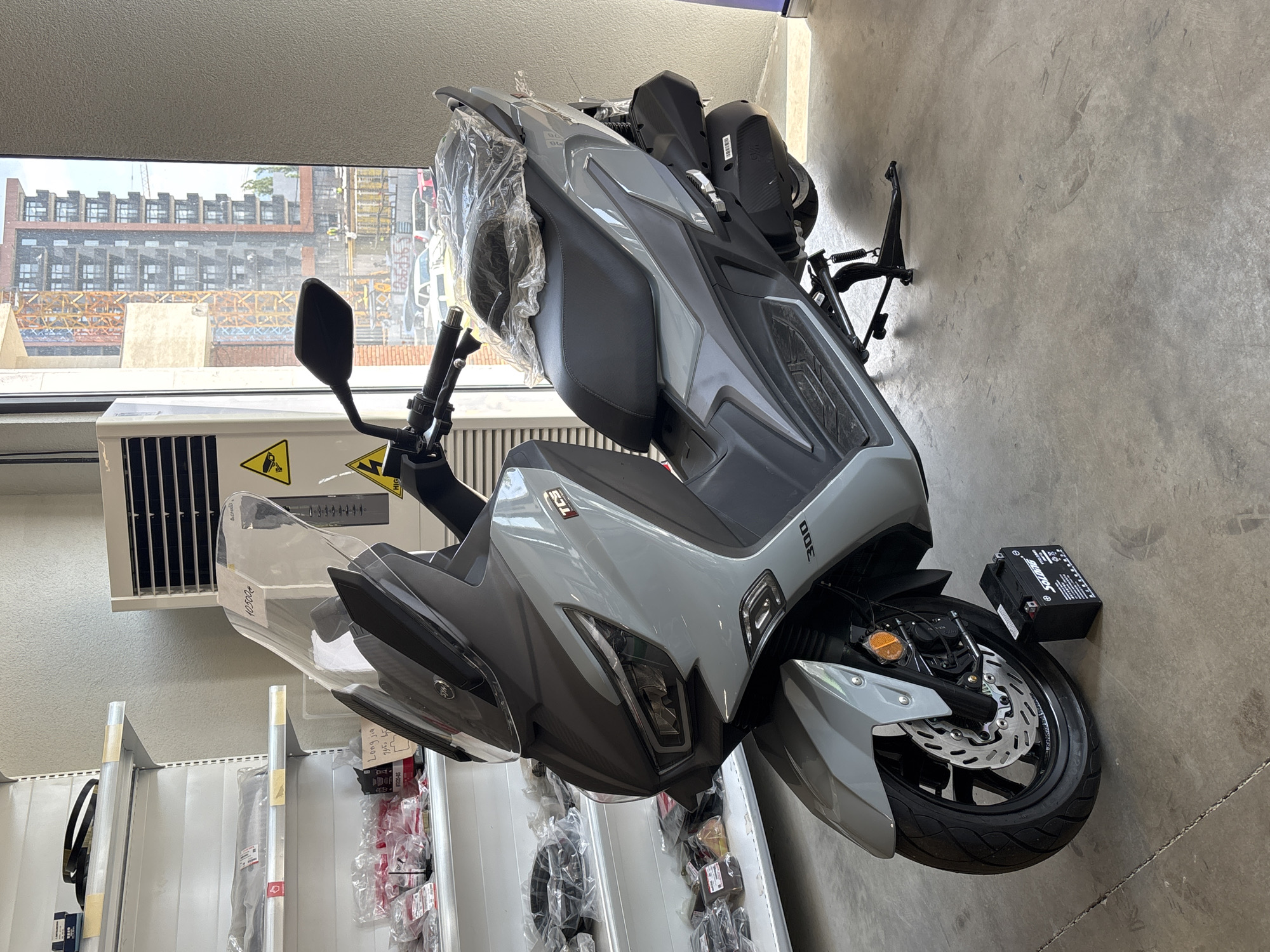 An indoor shot captures a light gray scooter with a clear windshield, parked on a concrete floor. The scooter features dark gray accents on the front, seat, and rear. A black seat is covered with clear plastic. A kickstand is deployed, holding the scooter upright. To the front left of the scooter, a black battery is placed on the floor. In the background, shelves stocked with packaged items are visible on the left. On the right, a large white air conditioning unit with yellow warning stickers is positioned near a window. Through the window, a city skyline with buildings under construction, a crane, and some vehicles can be seen. The overall environment suggests a retail or storage space, possibly a motorcycle dealership, located in Tbilisi, Georgia.