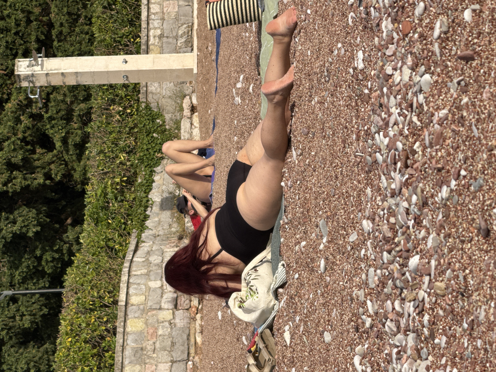 This image depicts a pebble beach scene in Sveti Stefan, Montenegro, under bright sunlight. The beach surface consists primarily of reddish-brown pebbles and small stones, with interspersed larger, lighter-colored stones.
In the foreground, a person with long, dark red hair is lying prone on a light green mat, facing away from the camera. They are wearing a black one-piece swimsuit. Their bare legs are extended towards the right side of the frame, with the soles of their feet visible. To their left, on the pebbles, a light-colored canvas tote bag with a green leafy design, a pair of tan sandals, and a red item are visible.
Further back and to the left, another individual is lying prone, with only their bare legs and lower torso visible on a blue mat. To the right of this person, closer to a stone wall, a third individual is also lying prone, wearing a dark cap and a red garment. Their bare legs are extended towards the center of the frame.
In the mid-ground, a light-colored concrete pillar functions as a beach shower structure, featuring two metal showerheads at the top and two lower spigots/faucets.
The background consists of a low stone wall, behind which dense green foliage, appearing as both manicured hedges and taller trees, is visible. A lamppost is partially visible among the trees on the left. All individuals appear to be sunbathing or relaxing on the beach.