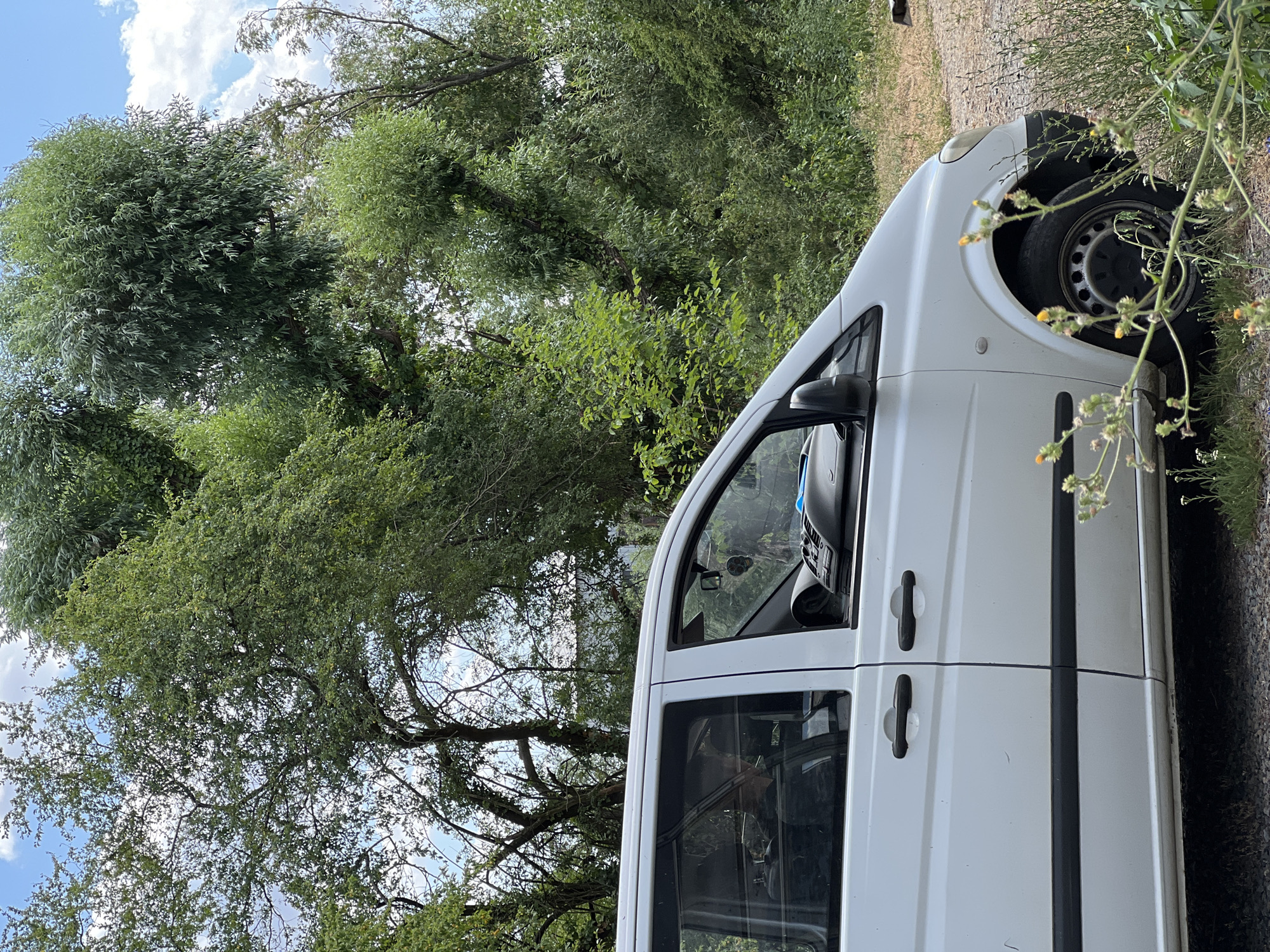 The image shows the side of a white van parked outdoors, with lush green trees and foliage surrounding it. The van is positioned at an angle, with its front wheel and part of the hood visible on the right side of the frame. The tires appear to be standard steel wheels. The driver's side door and rear passenger door of the van are visible, both with black door handles. The van's windows are dark, and reflections of the surrounding trees and sky can be seen in them.
The background is dominated by dense, leafy trees that create a natural, somewhat wild setting. The sky above is a mix of blue with fluffy white clouds. The ground next to the van appears to be a rough, gravelly surface with some dry grass and small wildflowers growing along the edge. The lighting suggests it is daytime, likely a sunny or partly cloudy day, given the brightness and the shadows.
The scene evokes a sense of being in a rural or natural environment, possibly a park, a forest edge, or a less-developed area. There are no people visible in the image. The overall impression is a peaceful, natural setting with a parked vehicle. No text is visible in the image. The location context provided is Lanžhot, Czechia.