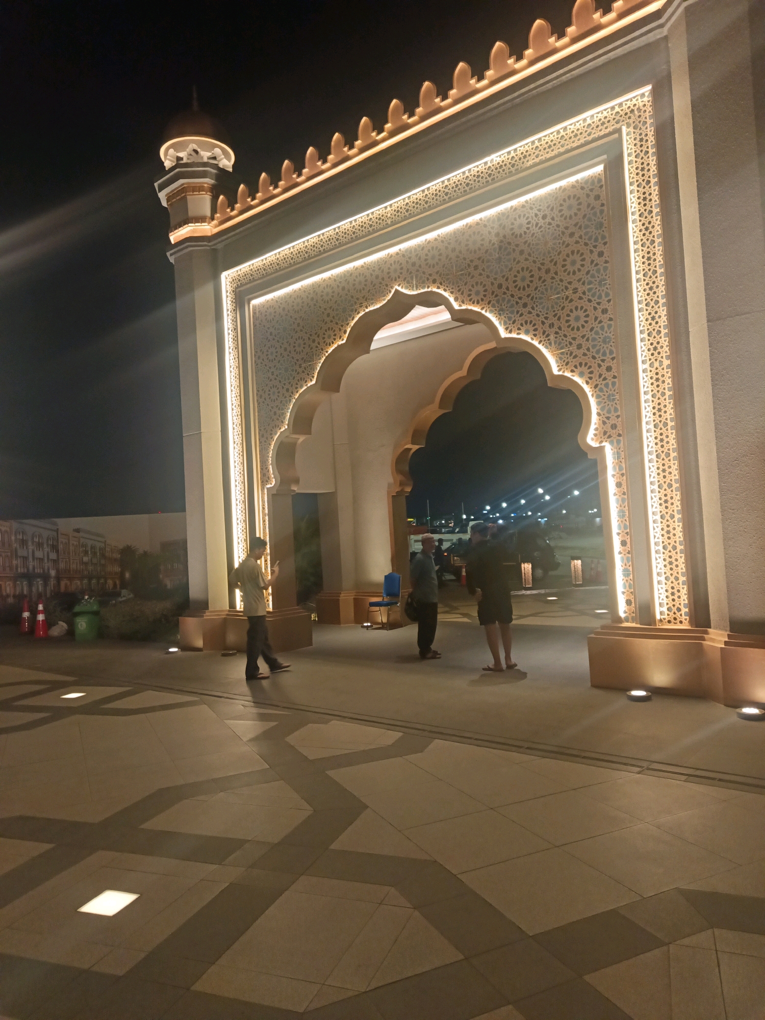 The image depicts the entrance to a building in North Jakarta, Indonesia, at night. The architecture features a large, ornate archway with Islamic-inspired geometric patterns and decorative lighting. Several individuals are visible near the entrance. One person stands to the left, looking at their phone, while two others are seen further inside the archway. The ground is paved with large tiles featuring a patterned design that creates shadows from the ambient lighting. In the background, glimpses of a city street with illuminated lights and some parked vehicles can be seen through the archway. The overall scene suggests an illuminated public space at dusk or night.
