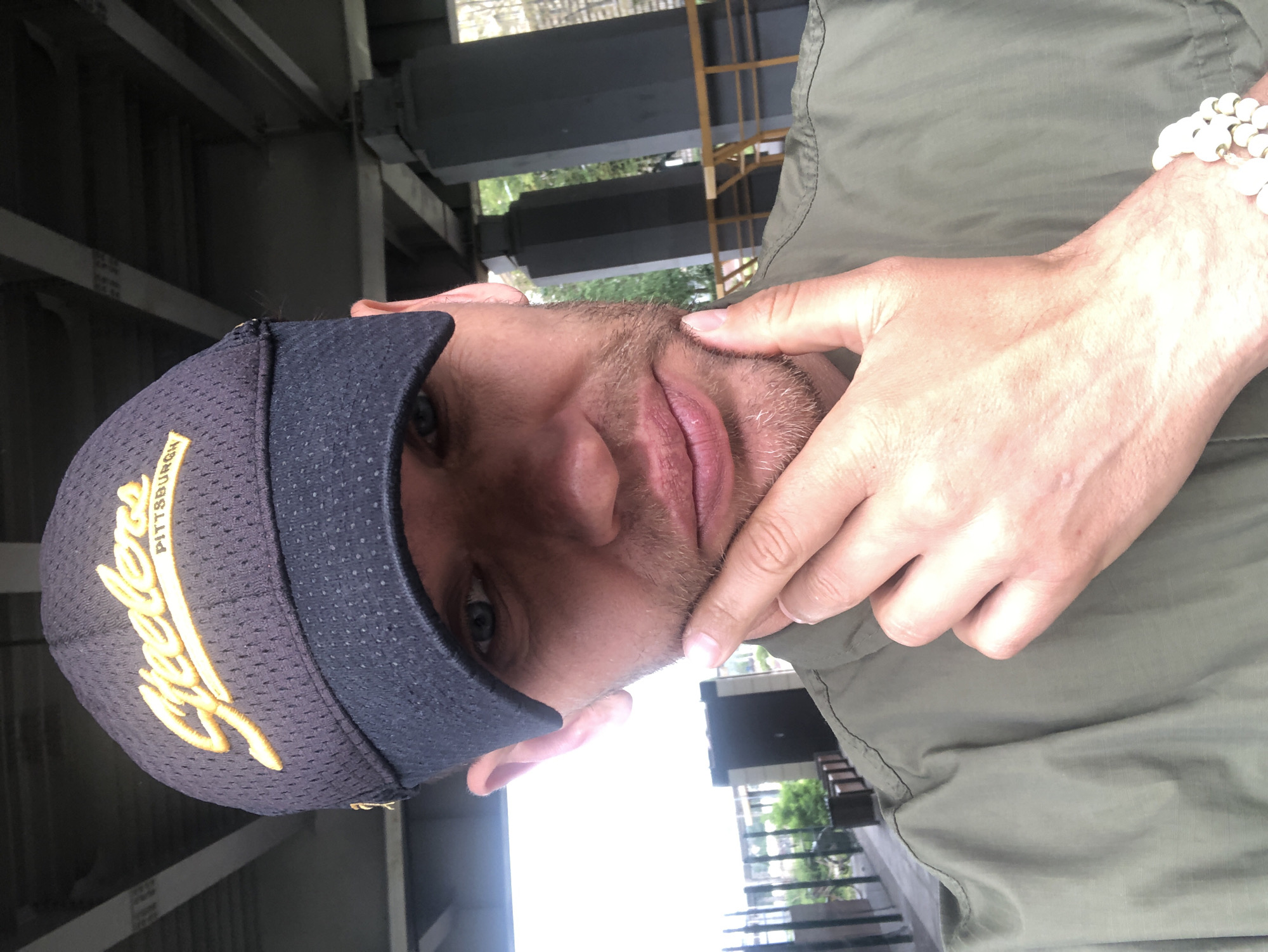 A man wearing a navy blue baseball cap with yellow lettering, an olive green jacket, and a white beaded bracelet on his left wrist is looking at the camera. He has stubble and is resting his chin on his right hand. The background shows large concrete pillars and yellow railings, suggesting an outdoor urban setting, possibly under a bridge or overpass. The environment appears to be Saint Petersburg, Russia, based on the architectural style of the pillars and railings which are common in Russian cityscapes.