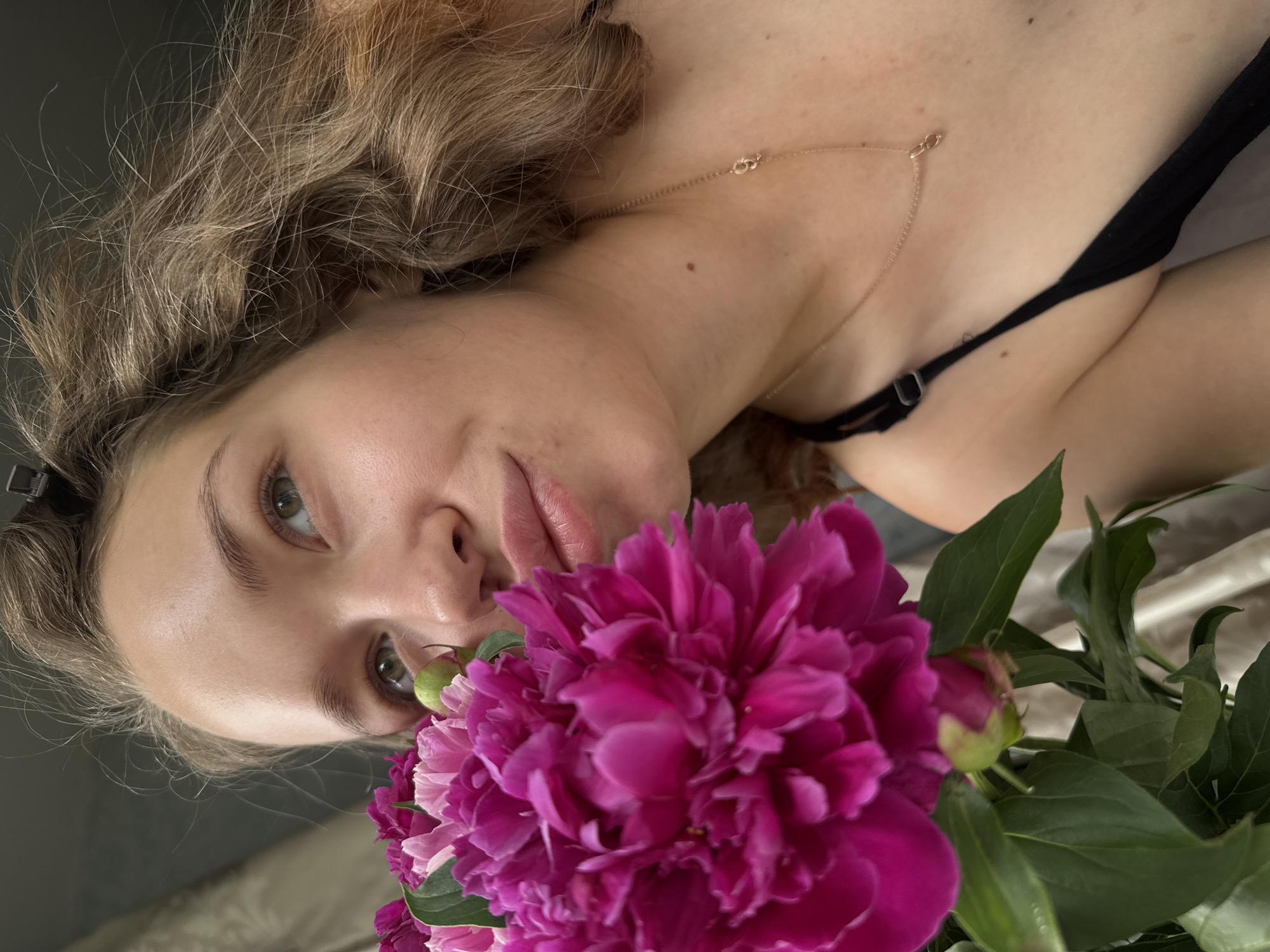 A young woman with blonde hair is holding a bouquet of pink peonies near her face. She is wearing a black top and a delicate gold necklace. The background is blurred, but appears to be an indoor setting with some soft fabric visible. There is no information about the location within the city of Chaikovsky, Russia.