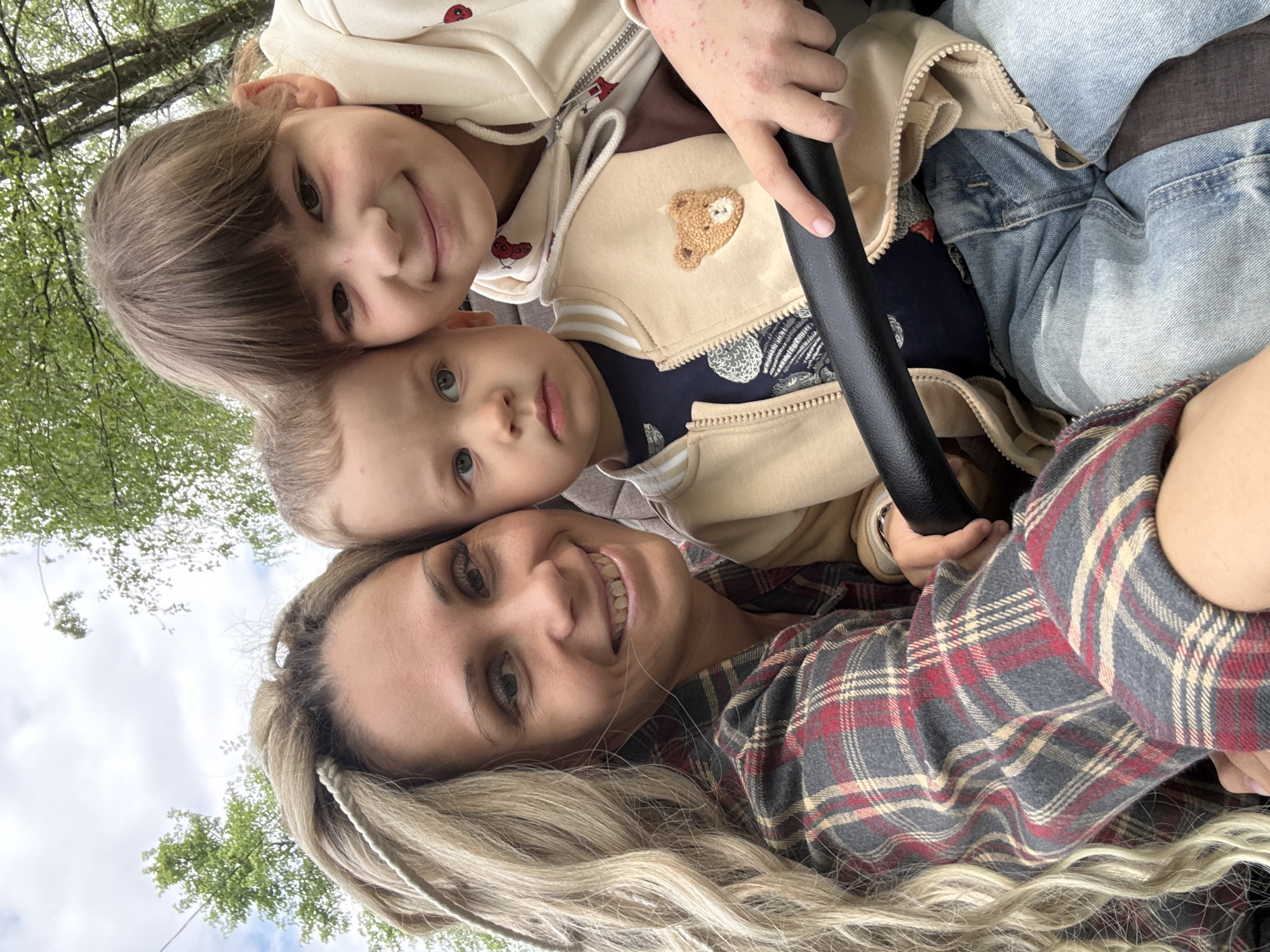 A selfie taken by a fair-skinned woman with long, braided blonde hair, wearing a red, gray, and beige plaid shirt. She is smiling at the camera. Above her are two fair-skinned children, a girl and a younger boy. The girl has brown hair and is wearing a light-colored hoodie with red mushroom designs. The boy has blue eyes and is wearing a beige jacket with a bear emblem. The girl's hand, showing some skin condition spots, is holding onto a black steering wheel. All three appear to be in a vehicle. The backdrop shows trees and sky, suggesting an outdoor setting. There is no specific information indicating the location as Rusanovo, Russia; this would require additional, specific features identifiable to that city.