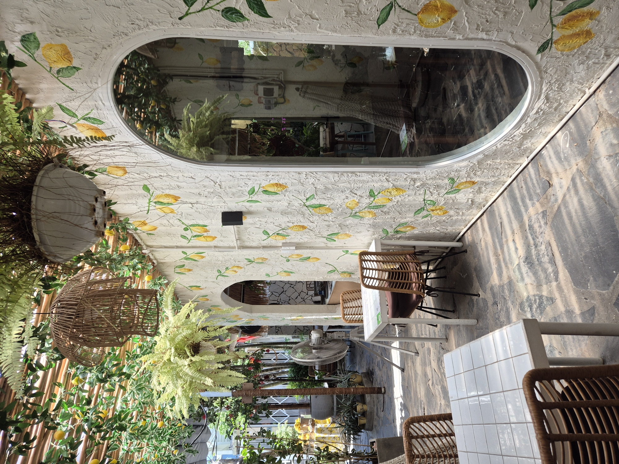 The image depicts the outdoor seating area of a cafe, likely located in the city of Đà Nẵng, Vietnam, as suggested by the presence of tropical plants and a generally warm, sunny ambiance often found in coastal Vietnamese cities. The cafe's exterior wall is adorned with a hand-painted mural of lemons and green leaves, adding a vibrant and whimsical touch. A large, oval-shaped window with a reflective surface dominates the right side of the wall, offering a glimpse into the cafe's interior.
The foreground features a white, tiled-top table with two brown wicker chairs, one of which is positioned in the lower left corner, partially obscuring the view. Another white table with two brown wicker chairs is visible in the middle ground, with a third chair and a table slightly behind it. A large industrial fan stands to the left of the tables, suggesting a need for cooling in the outdoor environment.
Above the seating area, a canopy made of bamboo slats is covered with lush green foliage and artificial lemons, creating a natural and shaded atmosphere. Hanging from the canopy are a large, woven wicker light fixture and a planter filled with cascading green ferns. In the background, the cafe's entrance can be seen through an arched opening, revealing more seating and decorative elements. The ground is paved with irregular stone slabs, and sunlight casts dappled shadows across the scene, indicating it's daytime. No people are present in the image.