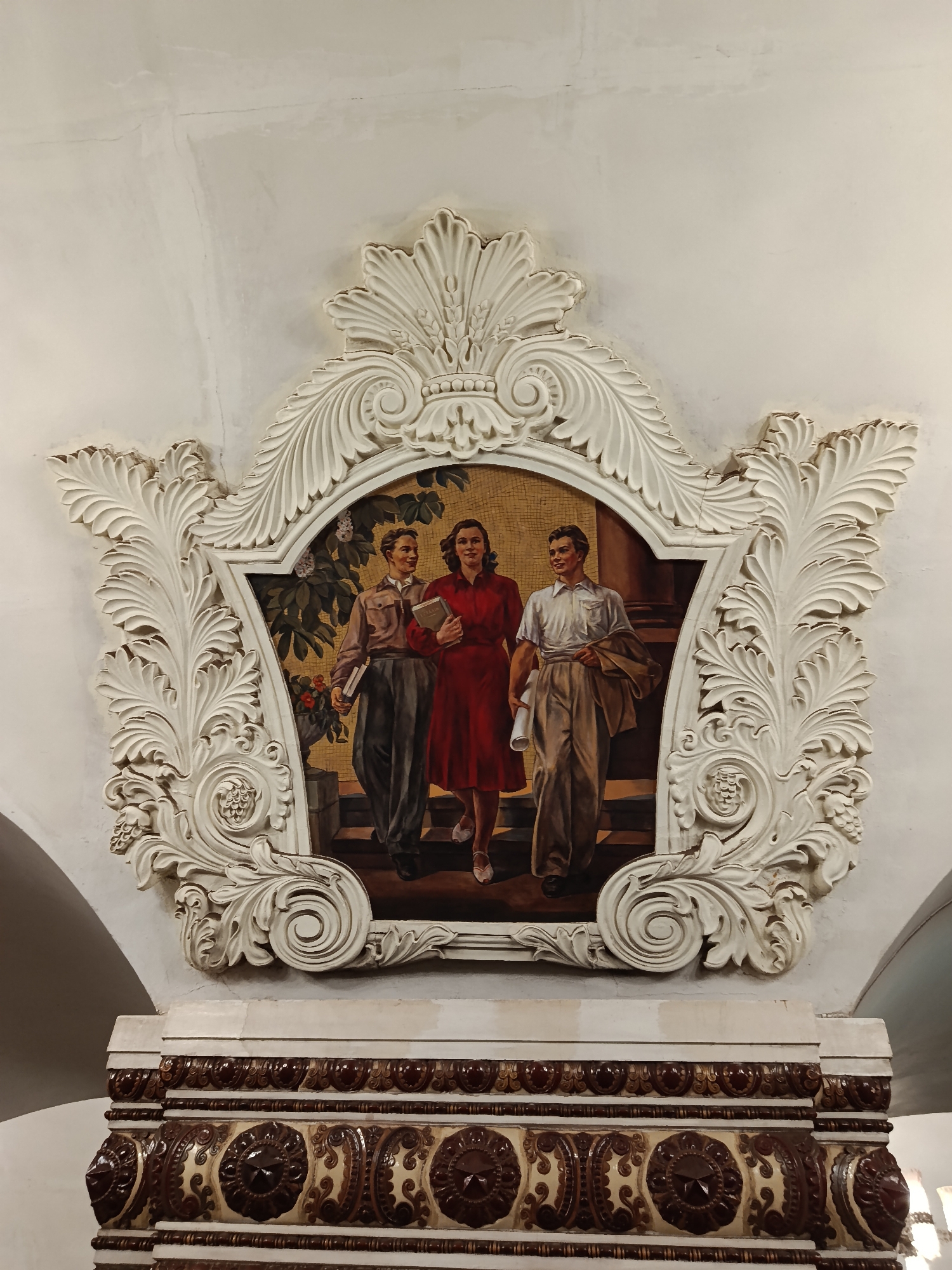 The image shows a mosaic in a decorative, white, ornate frame, depicting three figures. A woman in a red dress is in the center, holding a book. To her left is a man in a brown shirt and dark trousers, also holding a book. To her right is another man, in a white shirt and light trousers, carrying a jacket over his shoulder. They are on steps, with a background of foliage behind them. The mosaic is located above a decorative, brown and cream-colored tiled structure. This mosaic is located at the Kiyevskaya station on the Arbatsko-Pokrovskaya line of the Moscow Metro in Moscow, Russia.