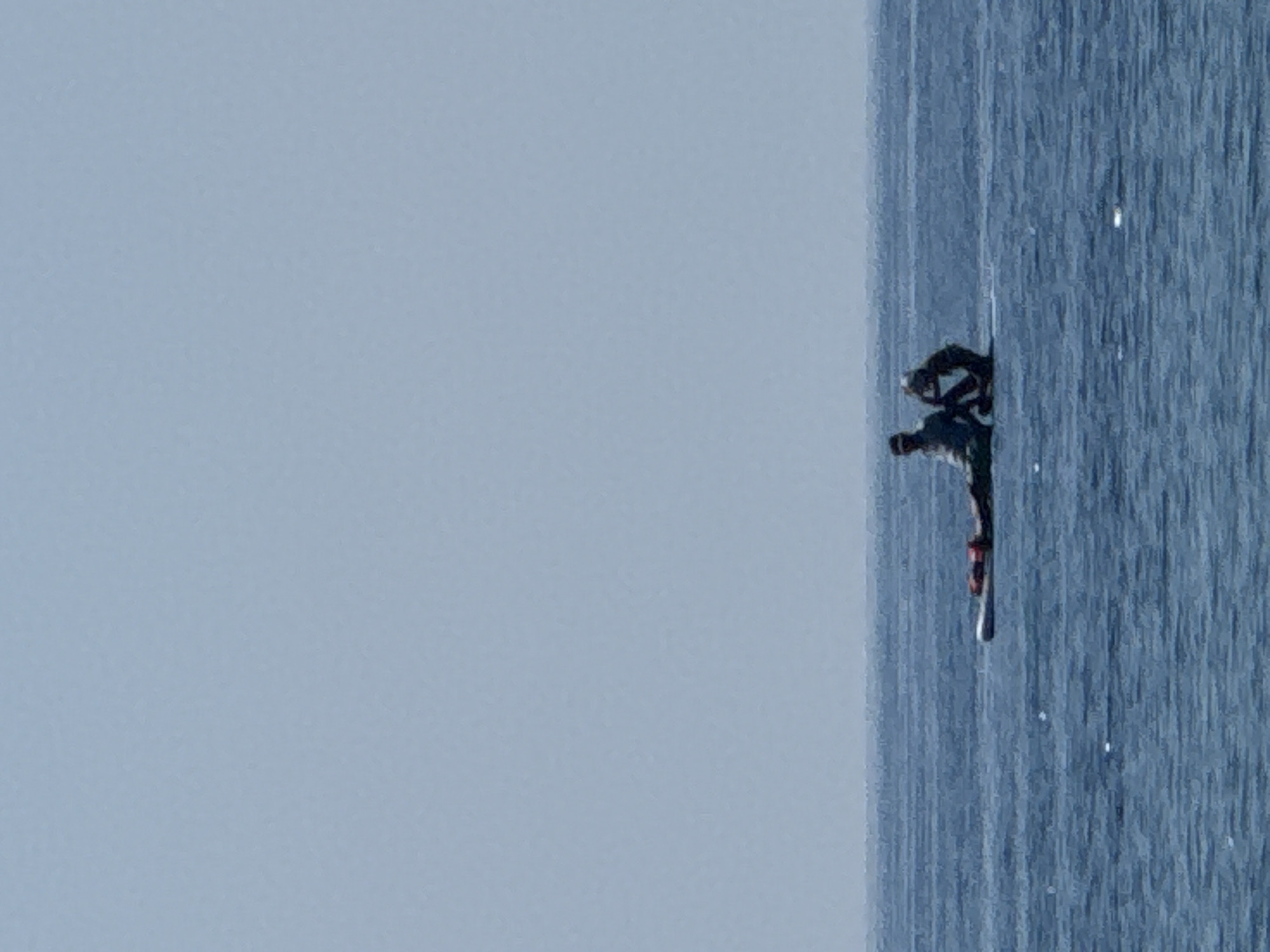 Two individuals are on a paddleboard in the sea, with the shoreline of Đurovići, Montenegro, visible in the distance. The person on the left is sitting with their legs extended, wearing a light-colored shirt and dark pants. The person on the right is kneeling and appears to be interacting with the other individual. Both are silhouetted against the bright sky. The water is a deep blue, with ripples indicating a calm sea. The sky is clear and light blue, meeting the sea at a distinct horizon line.