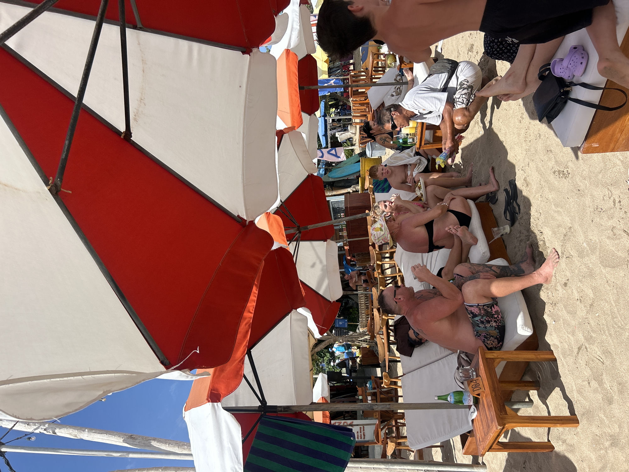 This is an eye-level medium shot of a beach scene in Kuta, Indonesia, under a cluster of large red and white umbrellas. In the foreground, a man with extensive tattoos on his arms and legs, wearing patterned swim trunks and sunglasses, is seated on a white lounge chair, holding a glass and looking towards the right. To his left, another man wearing swim trunks and a hat is seated and looking down at food on his lap. A woman in a black bikini is seated next to him, looking forward. Behind them, a young boy in a white towel is seated on a lounge chair, and a man in a light-colored shirt and patterned shorts is kneeling, holding a package and looking towards the seated group. In the middle ground, several other people are visible in the background, some seated at tables and others walking. There are lounge chairs, wooden tables, chairs, bottles of water, and umbrellas scattered across the sandy beach. The environment appears to be a beachside cafe or resort.