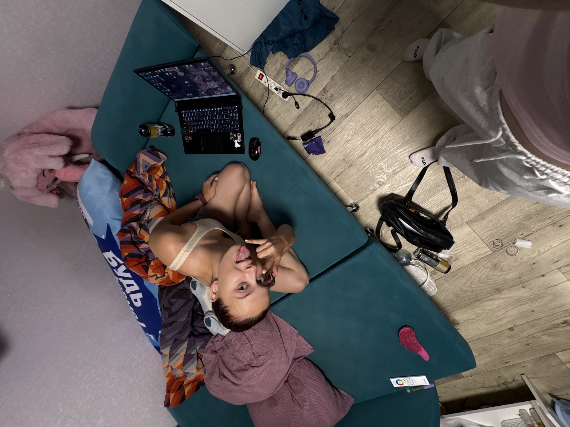 This image captures an indoor residential scene in Прикамский, Russia, taken from a high-angle, first-person perspective looking down into a room. A young woman with light skin and dark hair, pulled back, is positioned centrally on a multi-segment teal/green modular sofa. She is wearing a light-colored bralette and light blue socks, with her legs bent. Her left hand is raised in a gesture with fingers curled and thumb extended, while her tongue is slightly extended as she looks directly at the camera.
The sofa is draped with various colorful patterned blankets and throws in shades of orange, purple, grey, and black. A maroon/purple cushion and a blue cushion with partially visible white Cyrillic text are also on the sofa. A black laptop with an illuminated keyboard is open on the sofa, displaying a desktop background with a light-colored floral or abstract pattern. A black computer mouse rests beside the laptop. Next to the laptop, a yellow and black "FLASH UP" brand energy drink can with a straw is visible. On the left side of the sofa, a white rectangular remote control and a pink hairbrush are present. In the background, a large pink fluffy stuffed animal, resembling an elephant, rests against a light grey wall and the sofa.
The floor, covered with light-toned wooden laminate or plank flooring, contains several items. A white power strip with a red toggle switch is visible, plugged into an outlet, with multiple cables connected, including one leading to purple over-ear headphones and another to a coiled purple cable. A crumpled piece of blue fabric is near the power strip. A black bag with a strap, an empty amber-colored bottle or can, a pair of white flip-flops, a small white wrapper, and several metal rings are scattered on the floor. In the immediate foreground, the legs and feet of the person taking the photo are visible, wearing light grey sweatpants and white "FILA" branded socks. A partial view of a white cabinet or furniture piece is visible along the right edge of the image.
