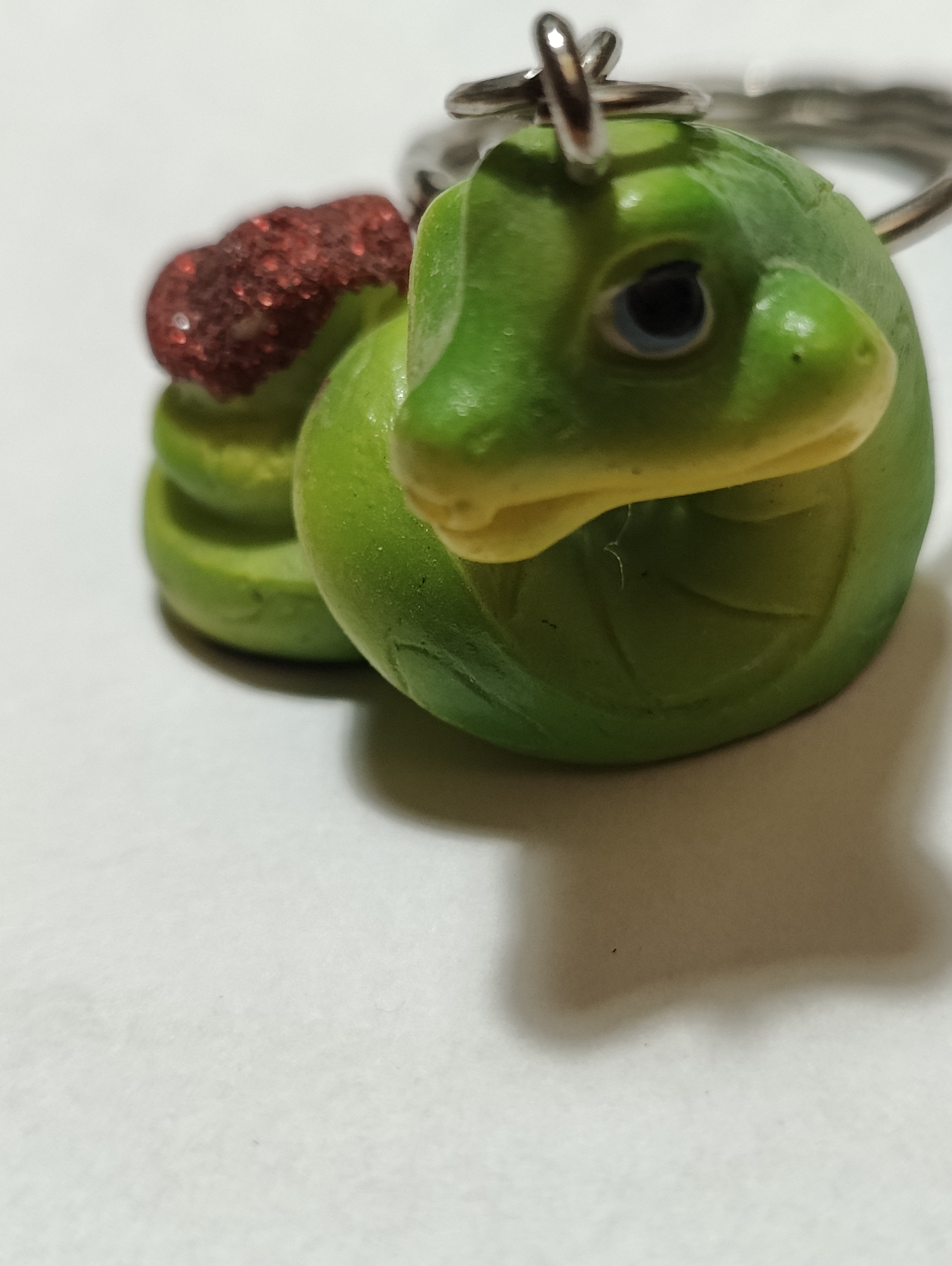 A close-up, overhead, shallow depth of field shot shows a green, turtle-shaped keychain with a red glittery, shell-like adornment. The turtle's head is facing forward, with its eyes visible. A silver keychain ring is attached to the top of the turtle's head. The background is a white, textured surface, with shadows cast by the keychain. There is no indication of a specific city or location.