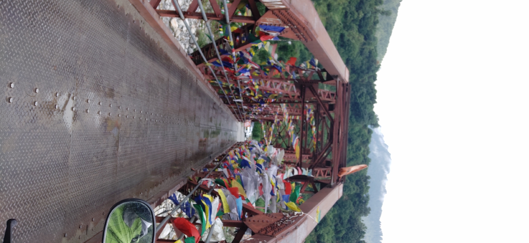 The image shows a perspective view of a rusted metal bridge in Kalath, India, adorned with numerous colorful prayer flags. The bridge's walkway is made of diamond plate metal and appears wet, with puddles reflecting the sky and surrounding elements. On either side of the walkway, metal railings are visible, and the bridge structure itself is a complex network of girders and supports.
Beyond the bridge, a lush green hillside is covered with trees, and in the distance, mist-shrouded mountains rise against a hazy white sky. The lower portion of the image captures a side-view mirror on the right, reflecting a person wearing a bright green jacket. The scene suggests a temperate, possibly slightly overcast day in a mountainous region.