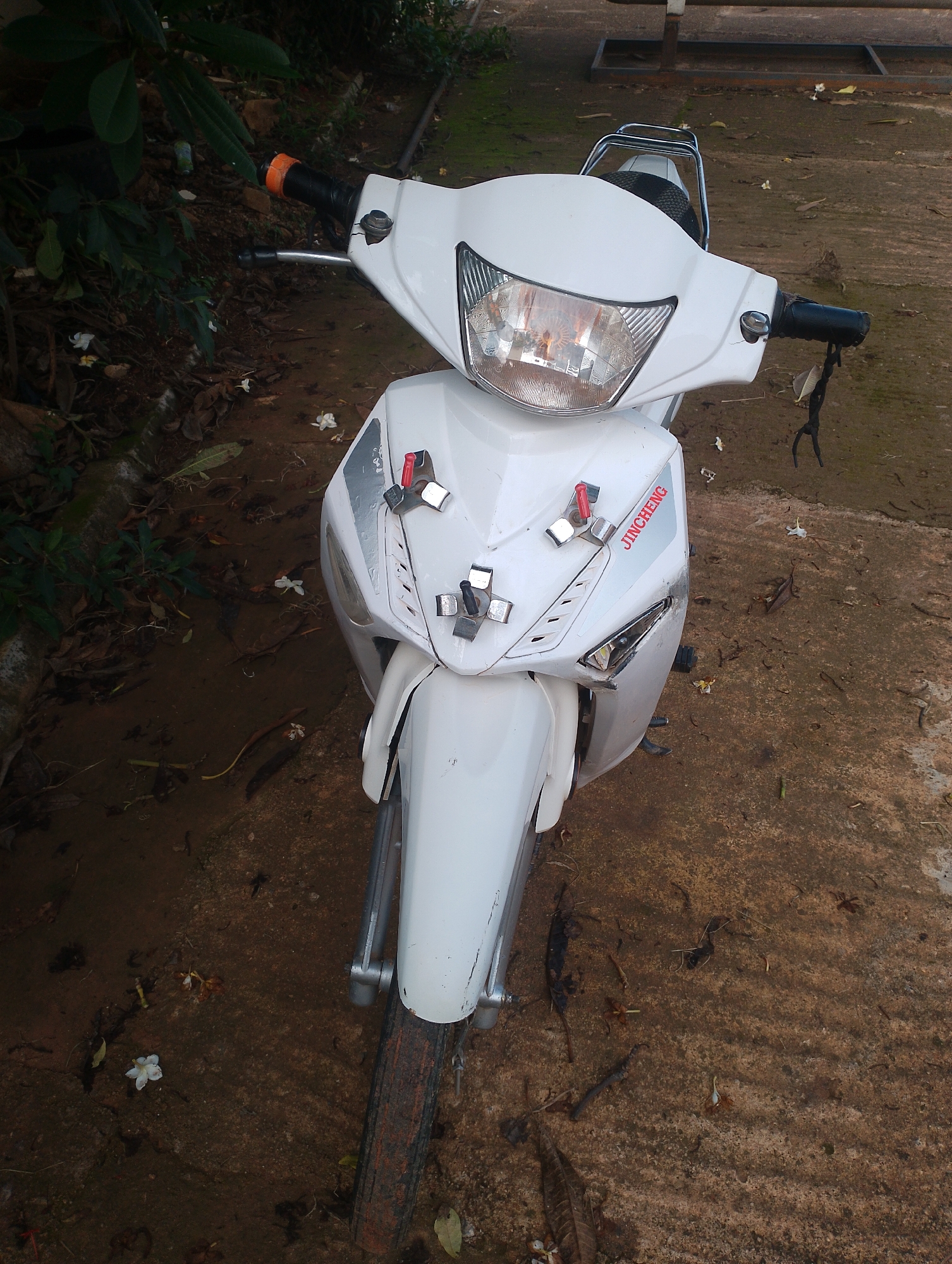 A white scooter is parked on a muddy and uneven ground, possibly a dirt path or an unpaved area in an outdoor setting. The scooter is facing the camera and its front is angled slightly to the right. The scooter has a shiny white exterior with some red accents on the front panel and a logo that reads "JINCHENG." The handlebars are black with an orange grip on the left side. A black cord or string hangs from the right handlebar, with a small, dry leaf attached to it. The ground around the scooter is a mixture of dark soil, small stones, and fallen leaves and petals, indicating an outdoor environment that may be somewhat neglected or naturally occurring. In the background, there are some dark green plants and foliage to the left, and a blurred metal structure that appears to be part of a bench or a frame to the right and top. The lighting suggests it is daytime, possibly late afternoon due to the soft light and shadows. The overall scene depicts a common urban or semi-urban environment, potentially within a residential area or a less maintained public space in Jos, Nigeria.