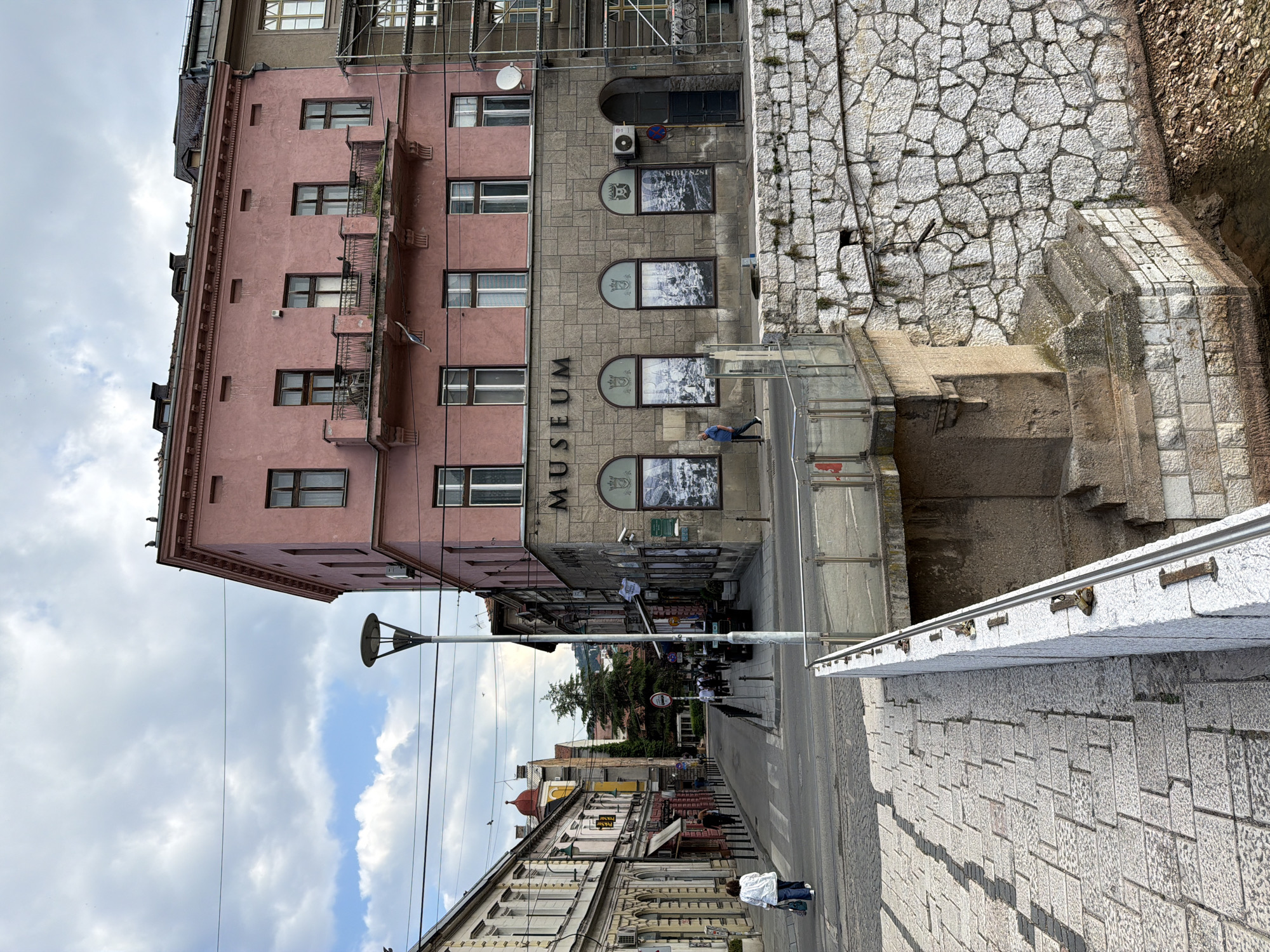 A street scene in Sarajevo, Bosnia and Herzegovina, is depicted from an elevated perspective looking down a cobblestone street lined with buildings. In the foreground, a stone-paved bridge or walkway with a white railing leads the eye into the scene. To the right, a textured stone wall forms the edge of what appears to be a canal or riverbed, with water and pebbles visible at the bottom.
Dominating the mid-ground is a pink-colored building with a distinct architectural style, featuring multiple windows and a balcony. Below the upper floors, the building's facade is made of stone with arched windows displaying framed images. The word "MUSEUM" is prominently displayed above these windows. A street lamp stands near the corner of this building.
The street itself is paved with cobblestones and appears to be in a city center. Several people are visible walking along the sidewalks or crossing the street. Vehicles are parked along the sides of the street, and traffic signs are present. The buildings lining the street exhibit various architectural styles and colors, suggesting an area with historical significance. Overhead, power lines crisscross the sky, and the sky is partly cloudy with a mix of blue sky and white clouds. To the far right, scaffolding indicates ongoing construction or renovation work on another building.