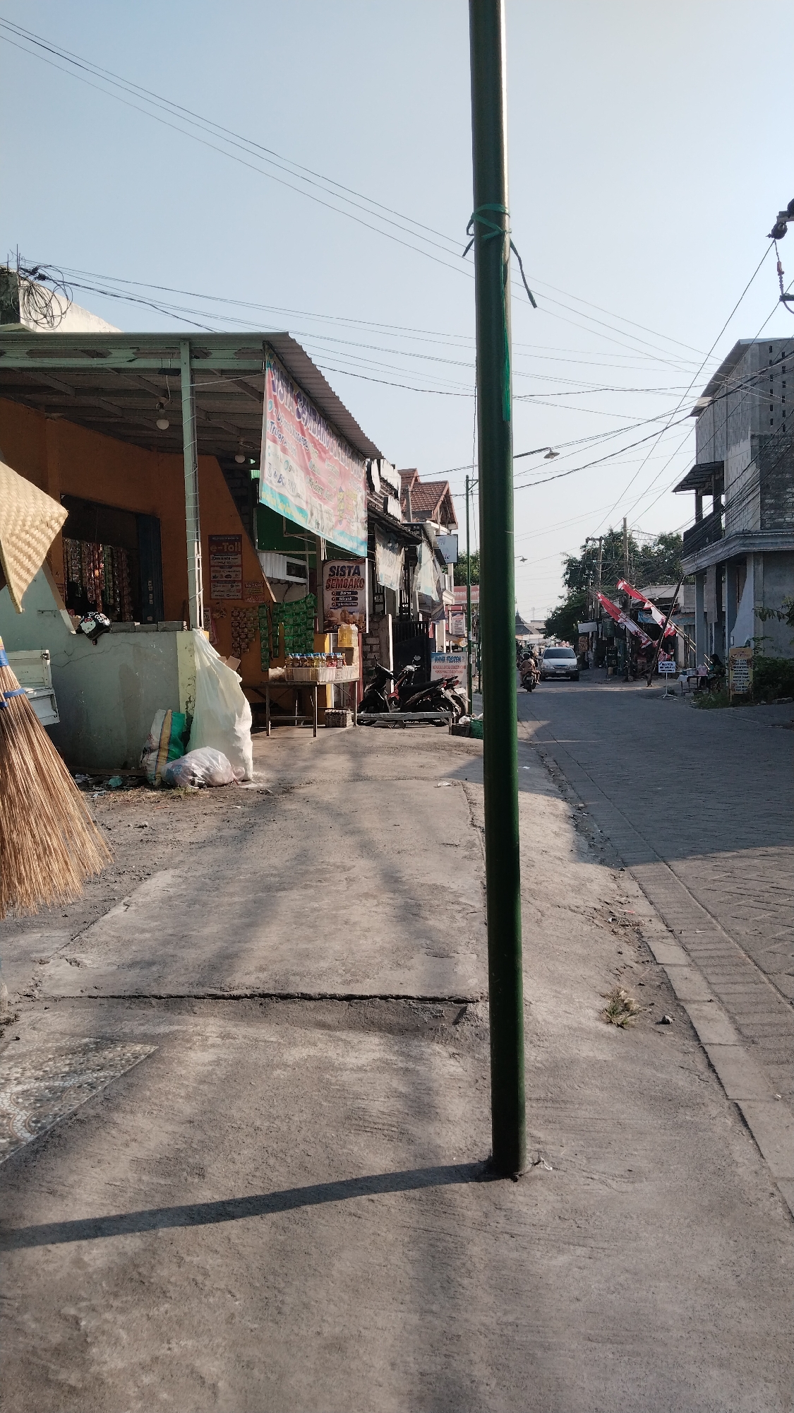 The image depicts a street scene in Peganden, Indonesia, under a clear sky. A prominent green pole stands in the foreground, casting a long shadow across the concrete pathway. To the left, a building with an orange facade is partially visible, featuring a covered walkway and a broom leaning against a wall. Various items, including sacks and what appears to be construction debris, are piled near the building. Further down the street, shops line both sides, with motorcycles parked in front of some. Red and white flags are displayed from one of the buildings. A white car is seen driving down the street, and several motorcycles are also present. The overall environment suggests a moderately developed urban or semi-urban area, with a mix of residential and commercial structures.