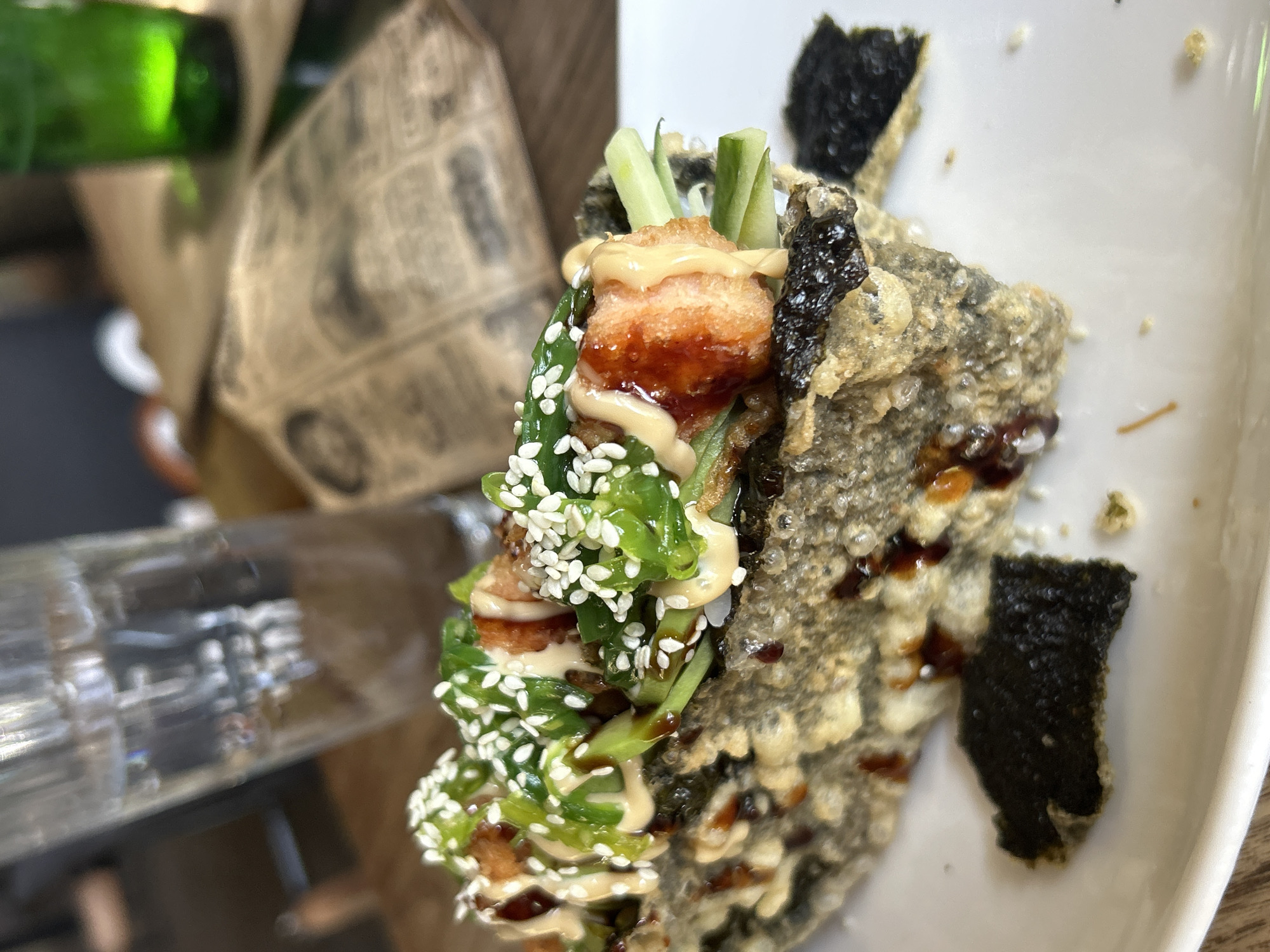 The image captures a close-up of a deep-fried sushi dish, likely a sushi taco or roll, presented on a white plate. The dish features a crispy, dark outer layer, possibly nori and tempura batter, with visible textures of frying. Inside, it is filled with chunks of what appears to be fried salmon, thin slices of cucumber, and a generous topping of chopped green onions and sesame seeds. A creamy sauce and a dark, glossy sauce are drizzled over the top. Several small pieces of dark, crispy nori are scattered on the plate around the main dish. In the background, a partially filled glass of water and a green glass are visible, along with a blurred, folded piece of what looks like a newspaper or menu. The scene is set on a wooden table. The presence of a newspaper and the style of the restaurant suggest an urban setting. While the image itself does not contain specific markers to pinpoint the location to Sofia, Bulgaria, such a dish would be commonly found in sushi restaurants within the city.