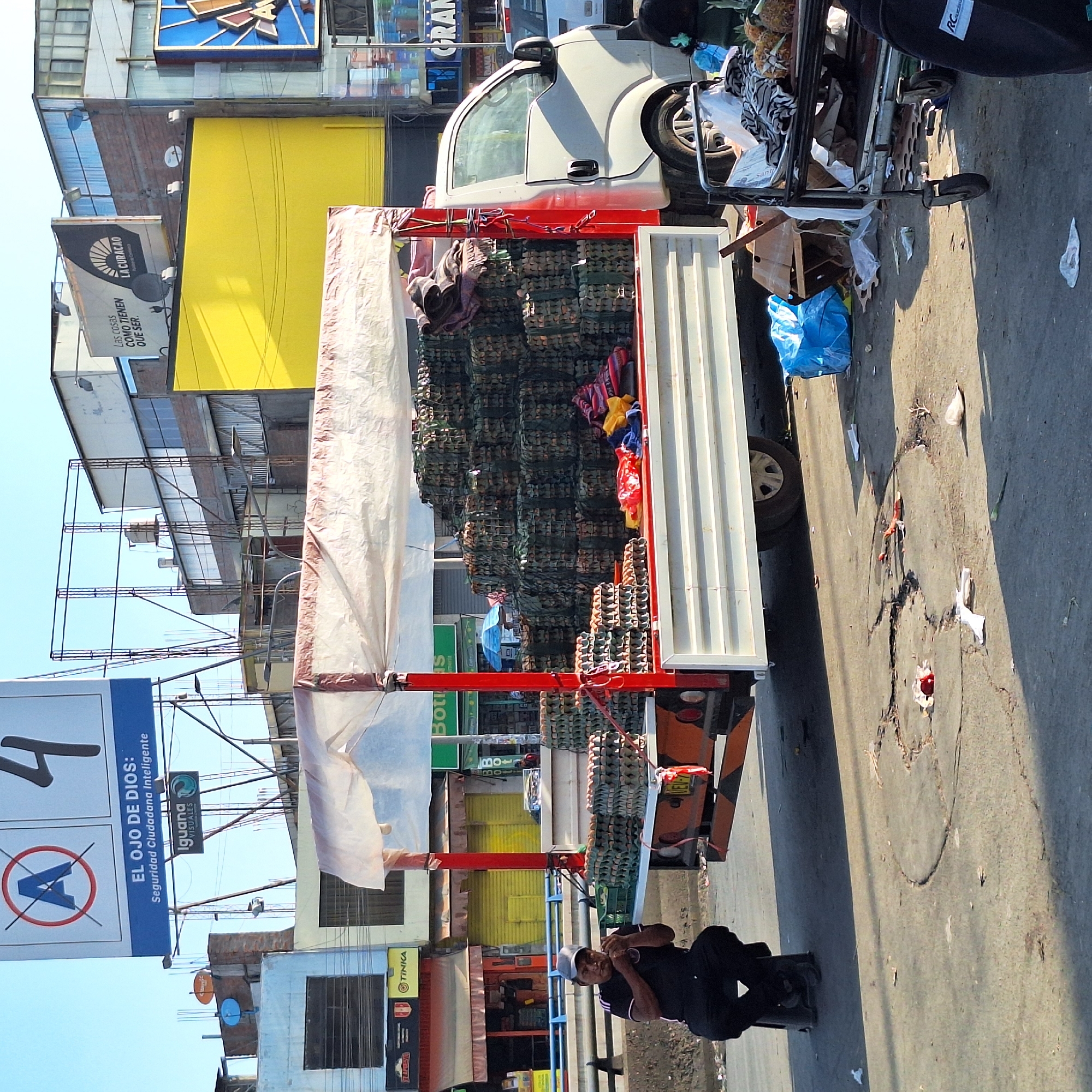 The image shows a street scene in José Luis Bustamante y Rivero, Peru. A white pickup truck is heavily loaded with stacked material in green netting. Next to the truck, a man in black clothing is seated on a small metallic chair. On the street, there is debris, a shopping cart, and a blue bag. Buildings and advertising signs are visible in the background. The signage includes advertisements for "LA CURACAO," "EL OJO DE DIOS," and "TINKA."