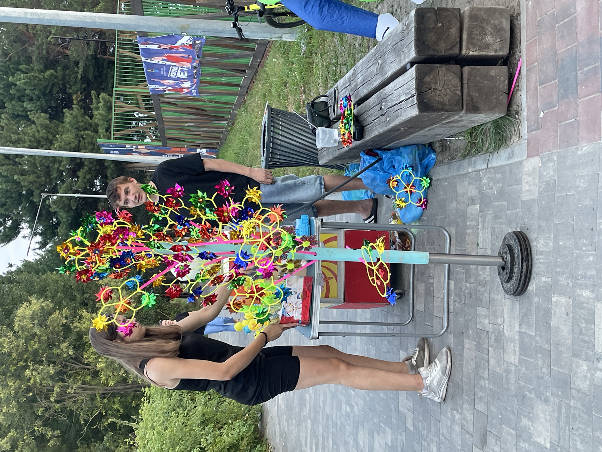 A young woman in a black tank top and shorts is seen selecting a colorful pinwheel from a display tree. Beside her, a young man in a black t-shirt and denim shorts observes. The display tree, adorned with numerous pinwheels, stands on a pavement. A metal table with a red base is set up next to the tree, holding a box of pinwheels. In the background, a green fence separates the area from a grassy field and trees. A trash can and a wooden bench are also visible. The scene appears to be outdoors, possibly in a park or public space in Surgut, Russia, given the context provided in the initial prompt.