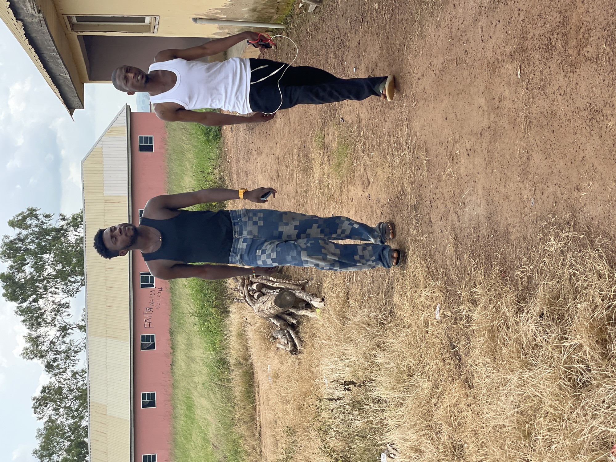 Two young men walk along a dirt path in Durumi, Nigeria. The man on the left wears a black tank top and patterned blue jeans, with sandals. He is looking towards the right. The man on the right wears a white tank top and black pants, with sneakers. He is looking forward and holding a red wired earphone set. Behind them, a long, single-story building with a pink facade and a light-colored metal roof is visible, with several windows. To the right of this building is a yellow-colored structure with a pitched metal roof. The foreground is a dry, grassy area next to the dirt path, with a pile of cut logs to the left. The sky is cloudy.