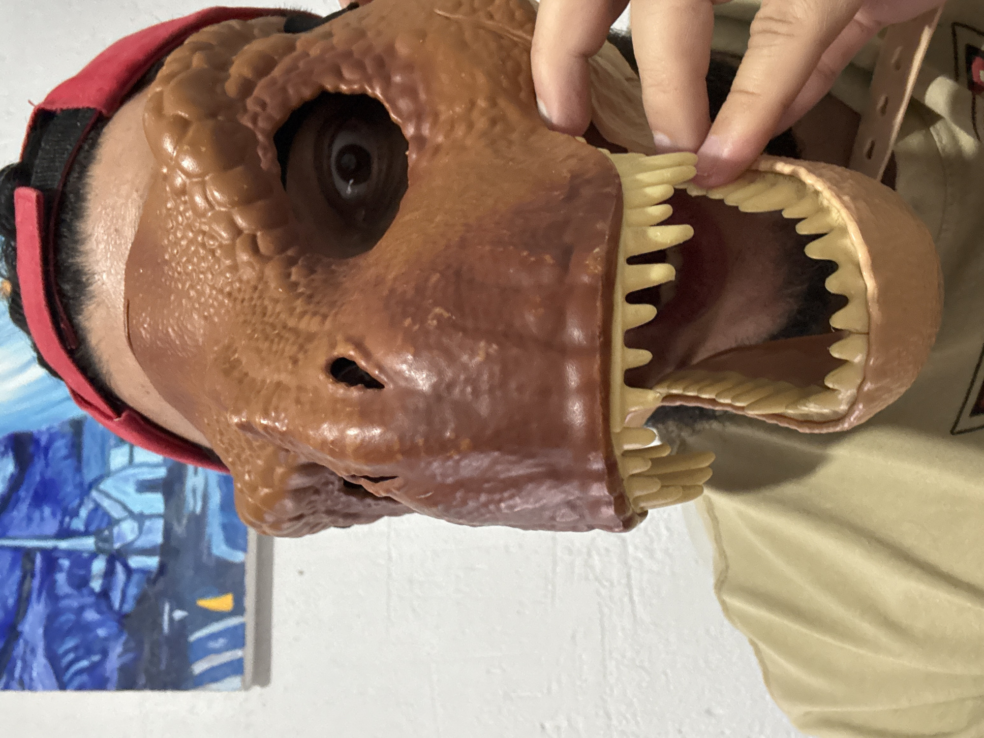 A person wearing a brown dinosaur mask with yellow teeth and a red baseball cap is shown from the chest up. The person's right eye is visible through an opening in the mask. A hand is reaching up to adjust the mask's jaw. In the background, a portion of a painting with a blue and white abstract landscape can be seen on a white brick wall. The setting is indoors.