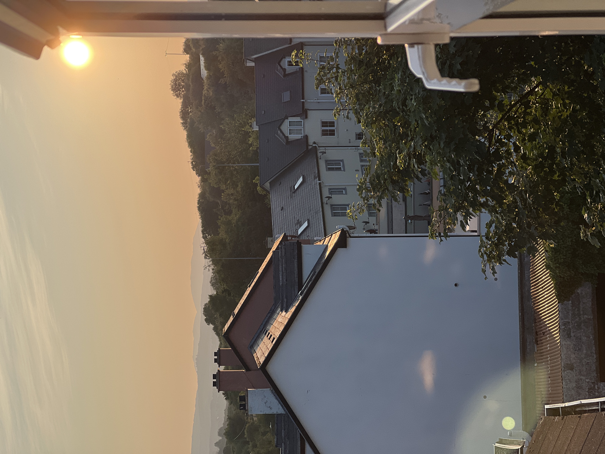 The image captures a sunset scene in the town of Mountcharles, Ireland. In the foreground, a white-walled building with a tiled roof is visible on the left, with another building featuring a reddish-brown roof and two chimneys behind it. Further back, a row of houses with dark tiled roofs and dormer windows is seen, partially obscured by lush green trees. The sky is a warm, hazy orange, with the setting sun a bright, prominent disc in the upper right quadrant, casting a golden glow on the scene. The landscape in the distance displays rolling hills, also softened by the atmospheric conditions. A window frame with a white handle is visible on the far right, suggesting the photo was taken from indoors.