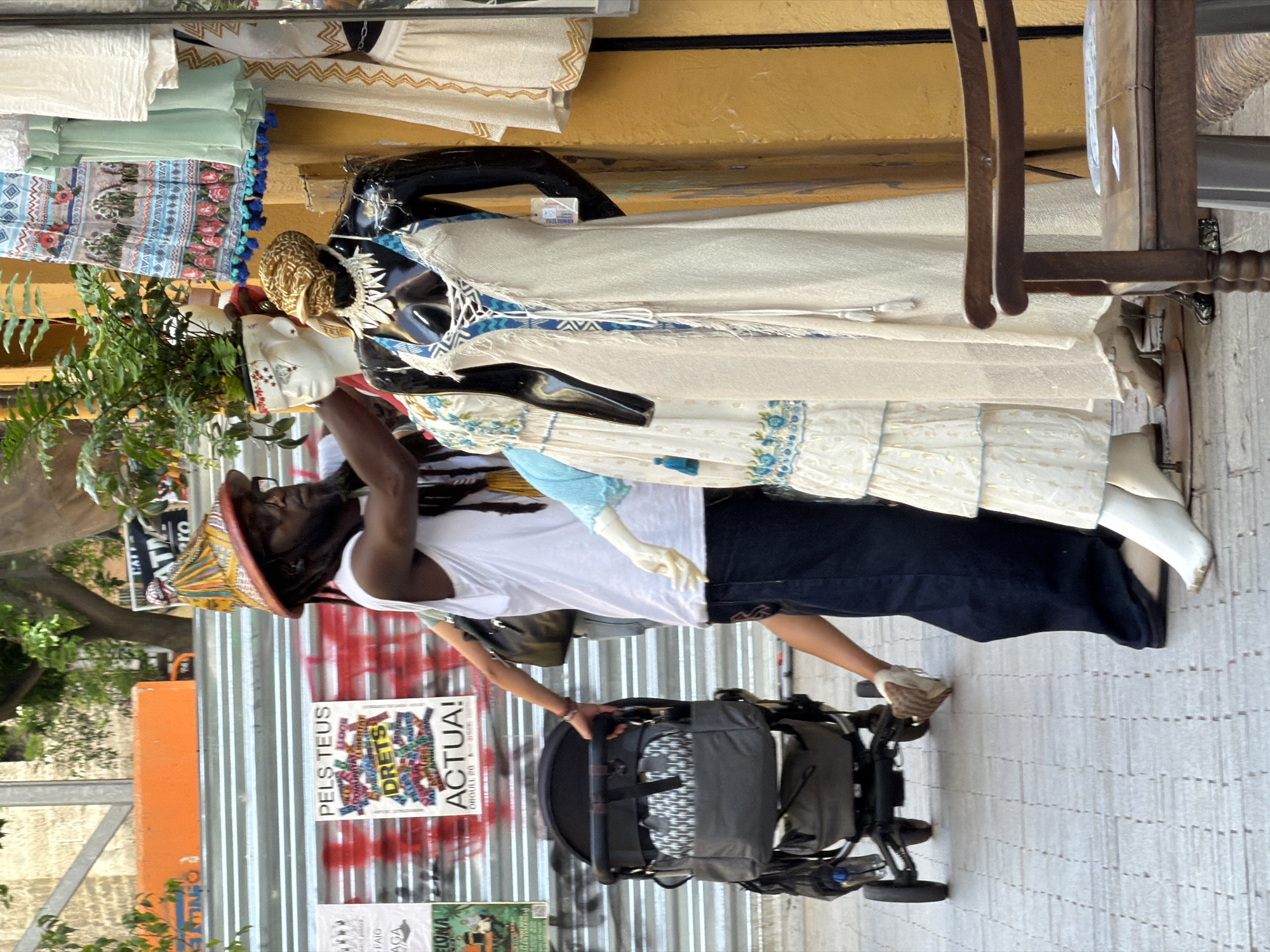 In an urban environment, a man with dark skin and dreadlocks, wearing a white tank top and dark pants, is pushing a dark gray baby stroller. He wears a distinctive, colorful, conical hat. To his right, a mannequin with black skin and a tan dress stands on display. Further to the right, another mannequin is partially visible, also wearing a tan dress. The background features a corrugated metal wall with graffiti and posters, including one that reads "PELS TEUS DRETS ACTUAL!". A plant in a white pot is visible behind the mannequin. In the foreground, a wooden chair with intricate carvings is positioned to the right of the mannequins. The ground is paved with grey interlocking blocks.