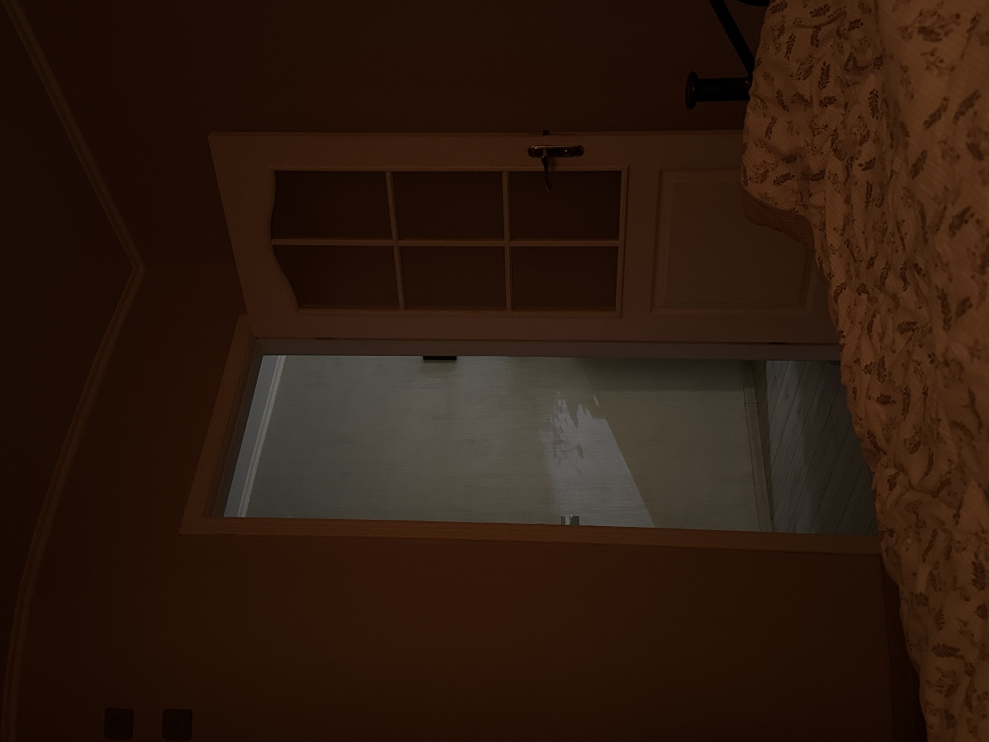 The image shows a partially opened white wooden cabinet door with a glass front divided into six rectangular panes. The door is slightly ajar, revealing the cabinet's interior, which appears to be a light-colored, possibly painted, surface. A metal latch is visible on the cabinet door. The wall surrounding the cabinet is a deep, muted brown. The lower portion of the image shows two electrical outlets set into the wall. Part of a light-colored patterned curtain or fabric is visible on the right side of the image.