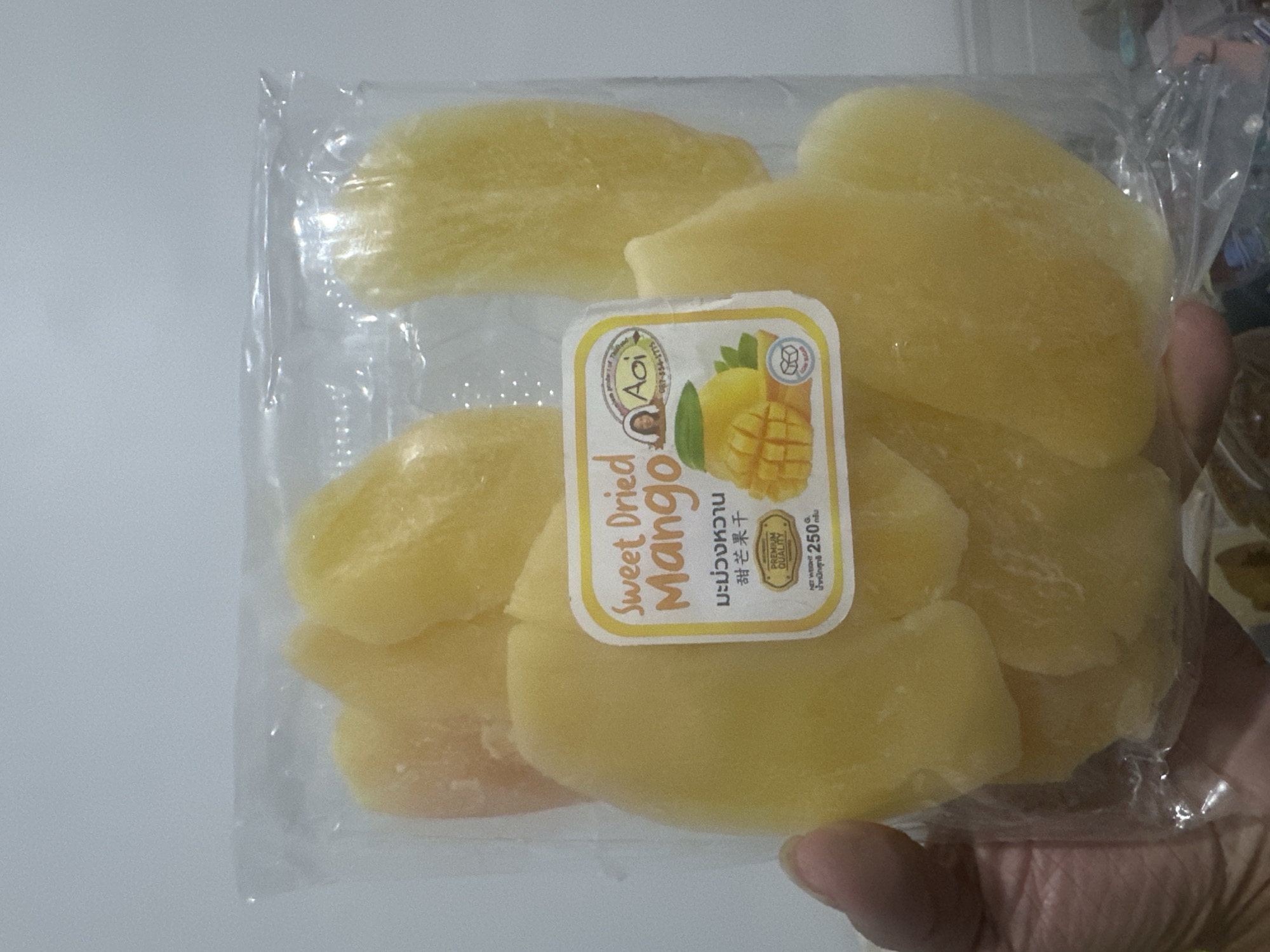The image displays a package of sweet dried mango slices. The packaging is clear plastic, revealing the yellow, translucent mango slices inside. A label is affixed to the front of the package. The label features the text "Sweet Dried Mango" in orange lettering, followed by Thai and Chinese characters. Below this, there is an illustration of a halved mango with segments cut out, alongside a green leaf. A small circular logo depicts a woman's profile and the text "Aoi." Further down, a "Premium Quality" seal is visible, and at the bottom, "NET WEIGHT 250 G." is printed. The package is being held by a person's hand, partially visible on the left side of the frame. The background is a plain, light-colored wall. This image does not contain any information that could be used to identify its location in the City of Binjai, Indonesia.