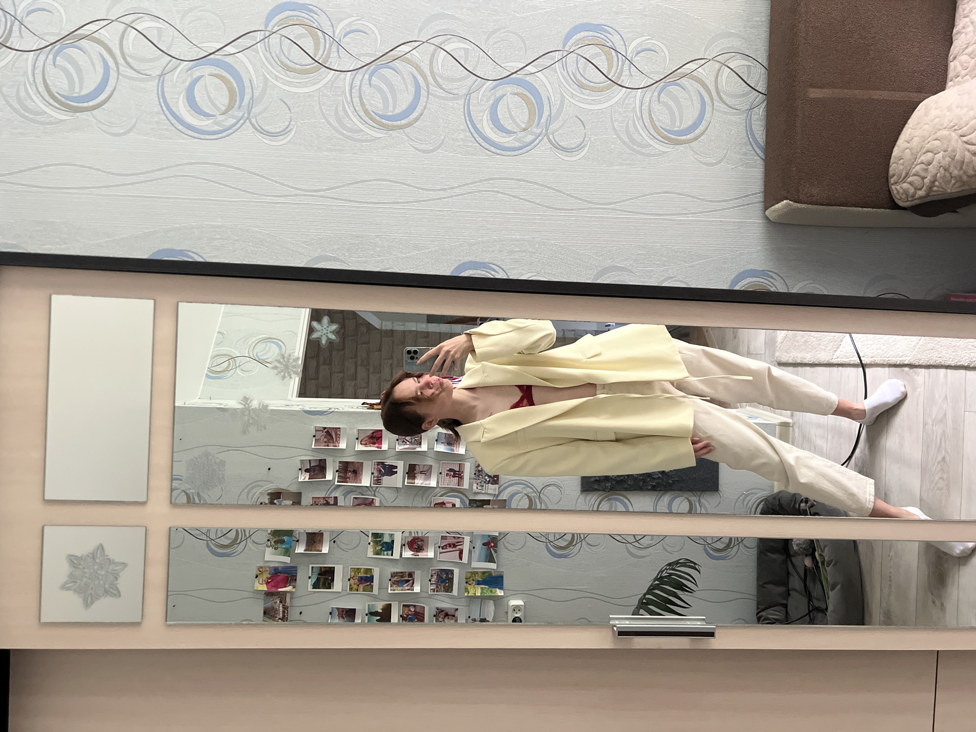 The image depicts an indoor residential scene in Chaikovsky, Russia, focused on a young female taking a mirror selfie. The central subject is a female in her late teens or early twenties, with light brown hair pulled back, holding a light-colored smartphone in her right hand to capture her reflection. She is wearing an open, oversized pale yellow blazer revealing a red bralette or bikini top, light-colored (cream or beige) drawstring casual pants, and white ankle socks. Her weight is predominantly on her left leg, with her right leg slightly forward.
She stands before a multi-paneled mirrored closet door with a light wooden frame. Numerous small, square instant-print photographs, likely personal memories, are affixed to the mirrored surfaces. Above the main mirror panels, two smaller, non-reflective panels are present, one featuring a raised snowflake or floral design.
To the right of the mirror, the wall is covered in light grey wallpaper with an abstract pattern of blue, grey, and light brown concentric circles and vertical wavy lines. To the left of the mirror, the wall is a plain light peach color. Reflections in the lower-left mirror panel show a dark grey pet bed containing green foliage and a small object, and part of a large green leafy plant. A white double electrical outlet is visible on the wall behind the reflected figure. The flooring consists of light wood laminate, partially covered by a cream-colored shaggy rug in the foreground. On the far right, the brown upholstered headboard and a cream-colored quilted pillow with a floral pattern of a bed are visible. Several decorative snowflake cutouts are taped to various reflective surfaces in the room.