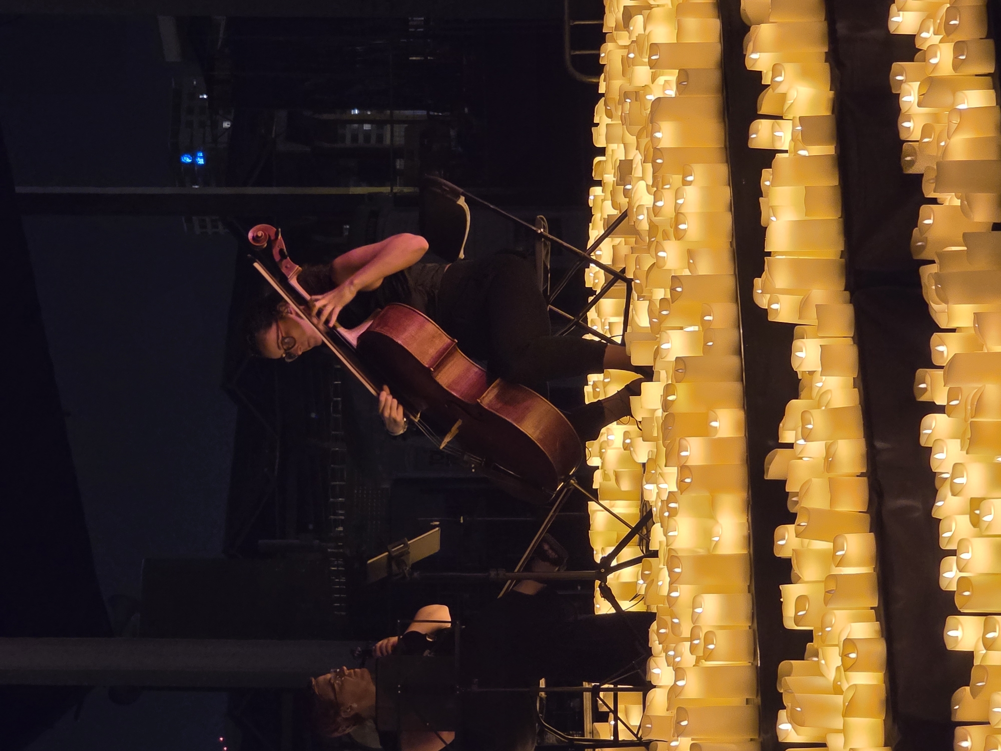 The image shows two women playing instruments in front of a large display of lit candles. The woman on the left is wearing glasses and a dark top, and she appears to be playing a string instrument that is out of frame. She is seated on a chair and looking towards the instrument. The woman on the right is playing a cello, and she is also seated on a chair. She is wearing glasses and a dark sleeveless top. The cello is a large wooden instrument with a rich brown color. The candles are arranged in rows on tiered platforms, and they are emitting a warm, yellowish glow. The background is dark, suggesting that the event is taking place at night. In the distance, there are faint outlines of buildings, some of which are illuminated.
Based on the context provided, the image depicts an event in the city of Sant Adrià de Besòs, Spain. The scene is outdoors, likely in a public space, given the presence of buildings in the background and the overall scale of the candle display. The atmosphere is serene and contemplative, accentuated by the soft lighting of the candles and the quiet activity of the musicians. The event appears to be a performance or a commemoration, given the formal setting and the presence of multiple candles, often used for memorial services or festive occasions. The composition of the image, with the musicians in the foreground and the city lights in the background, suggests a blend of artistic performance and urban environment.