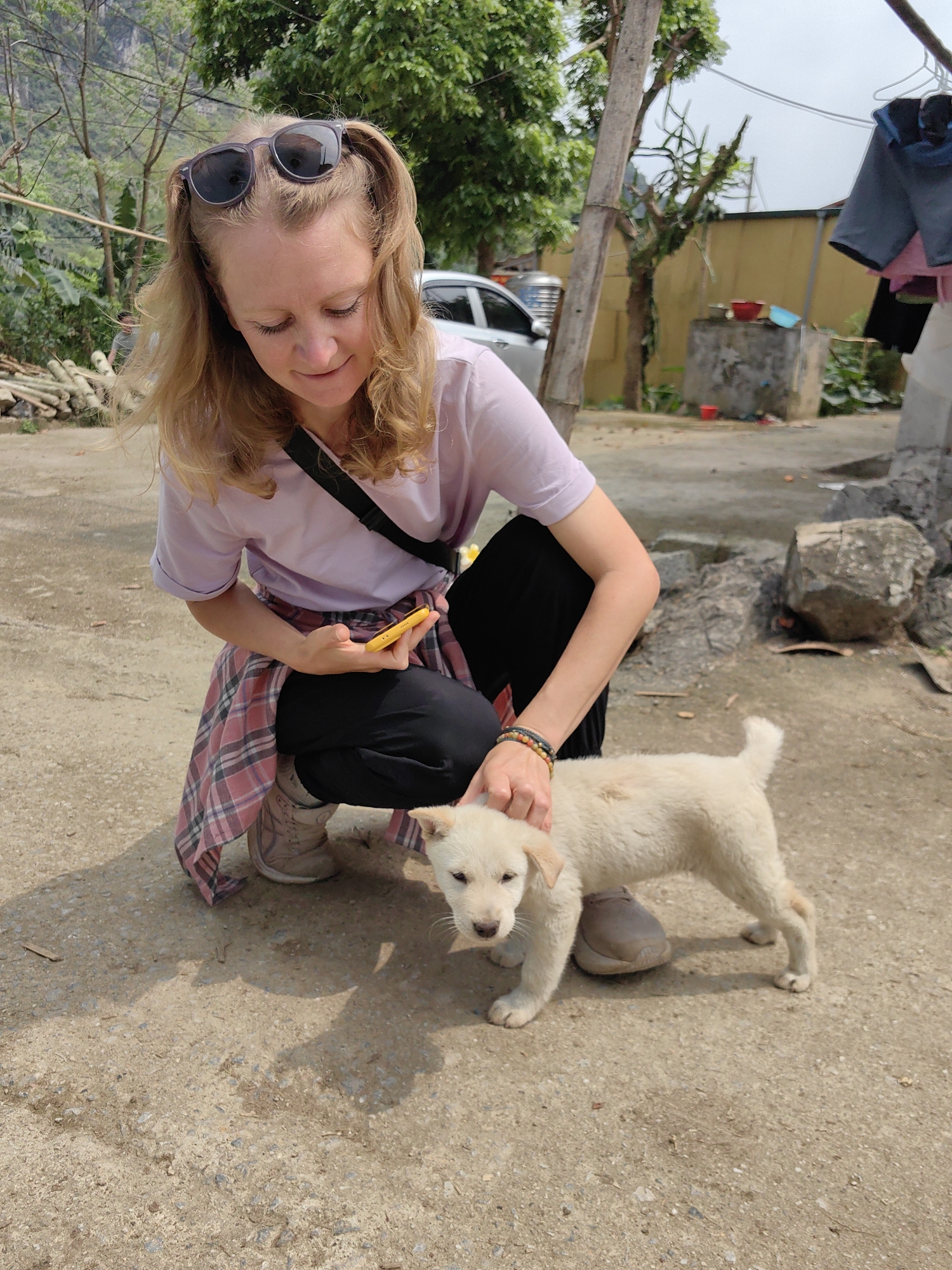 A fair-skinned woman is crouched down, petting a small, light-colored puppy on a paved surface. The woman is wearing a light purple t-shirt, black pants, and tan shoes. A pink and grey plaid shirt is tied around her waist, and she has a black strap across her shoulder. Sunglasses are perched on top of her blonde hair. She is holding a yellow smartphone in her left hand. The puppy stands in front of her, looking towards the camera. The background includes trees, a utility pole, a parked silver car, a concrete structure, and clothes hanging on a line. The location is Xã Đàm Thủy, Vietnam.